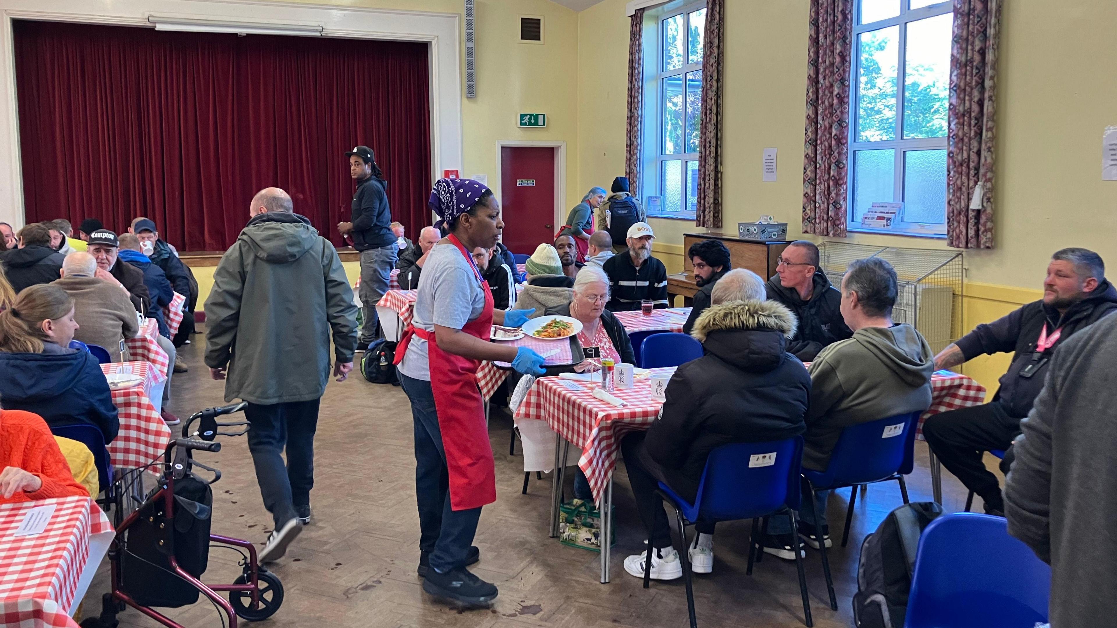 People seated down on table and chairs with a red and white table cloth and volunteers serving food in a tray