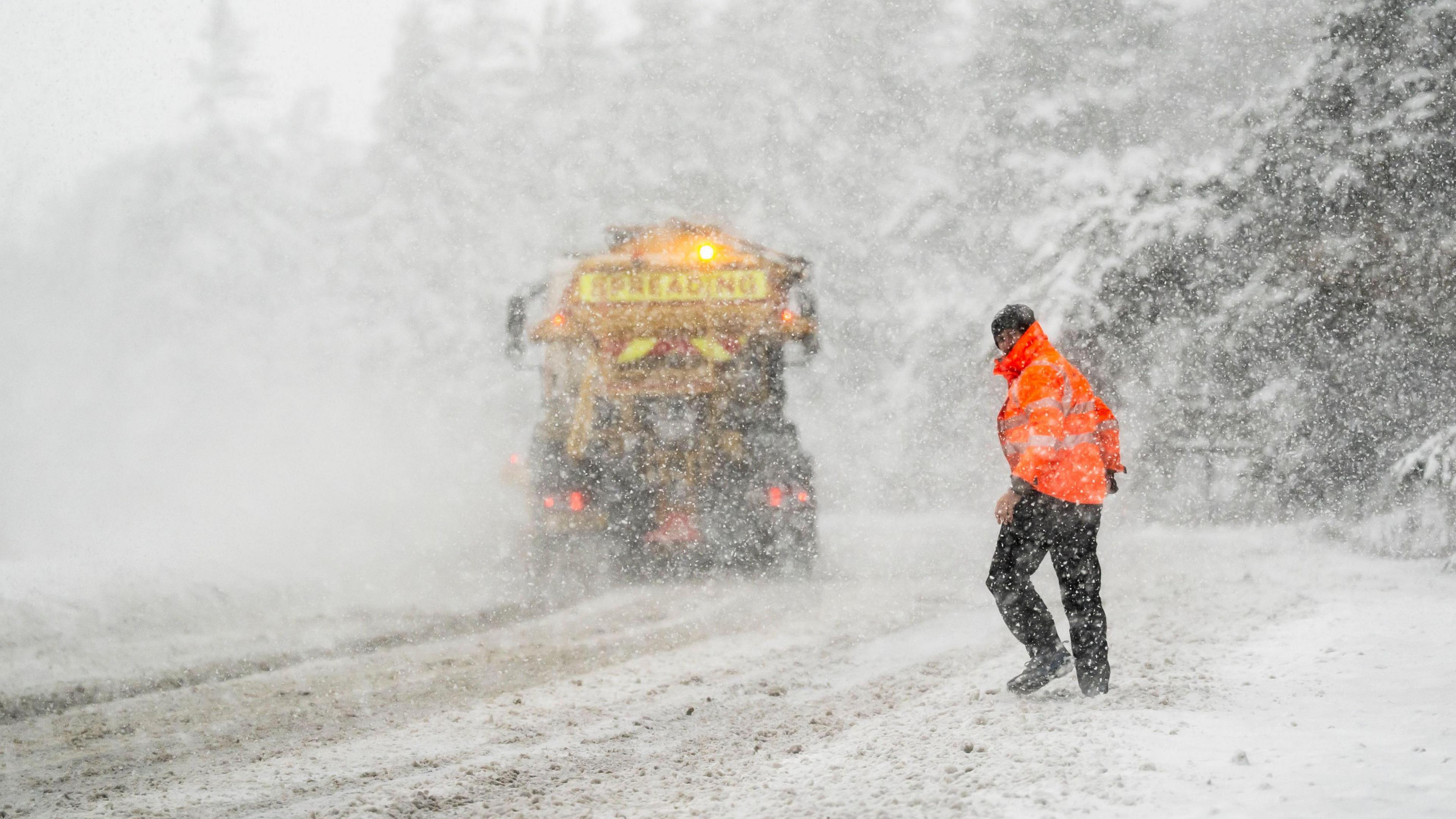 A National Highways employee on the A169 in the North York Moors walking on a snow covered road where a gritting lorry is also operating.  There is heavy snow falling