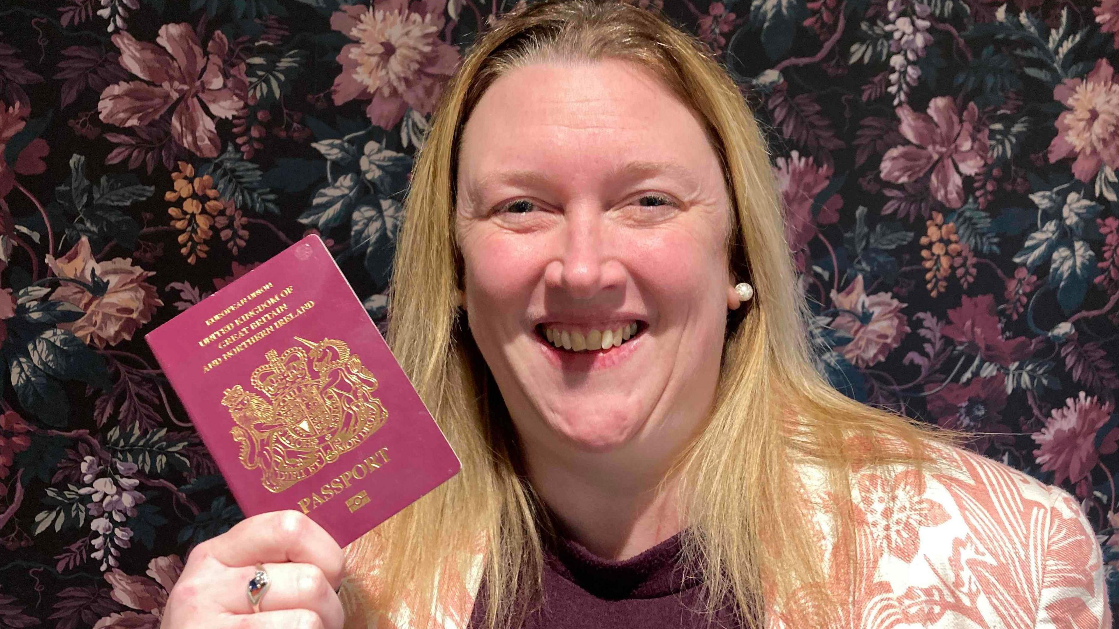 Rachel Bickford is holding up a passport. She has blonde hair, she is smiling and there is floral wallpaper behind her.