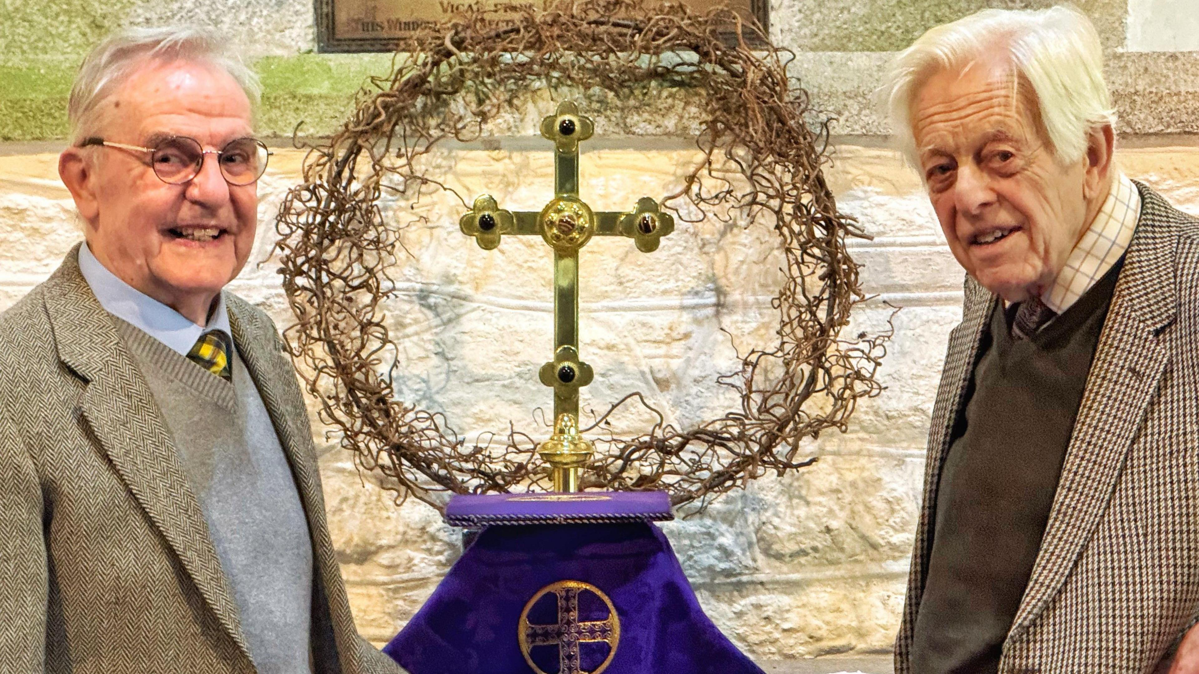 Two older men smiling at the camera with the brass cross surrounded by a willow wreath between them on the church altar 