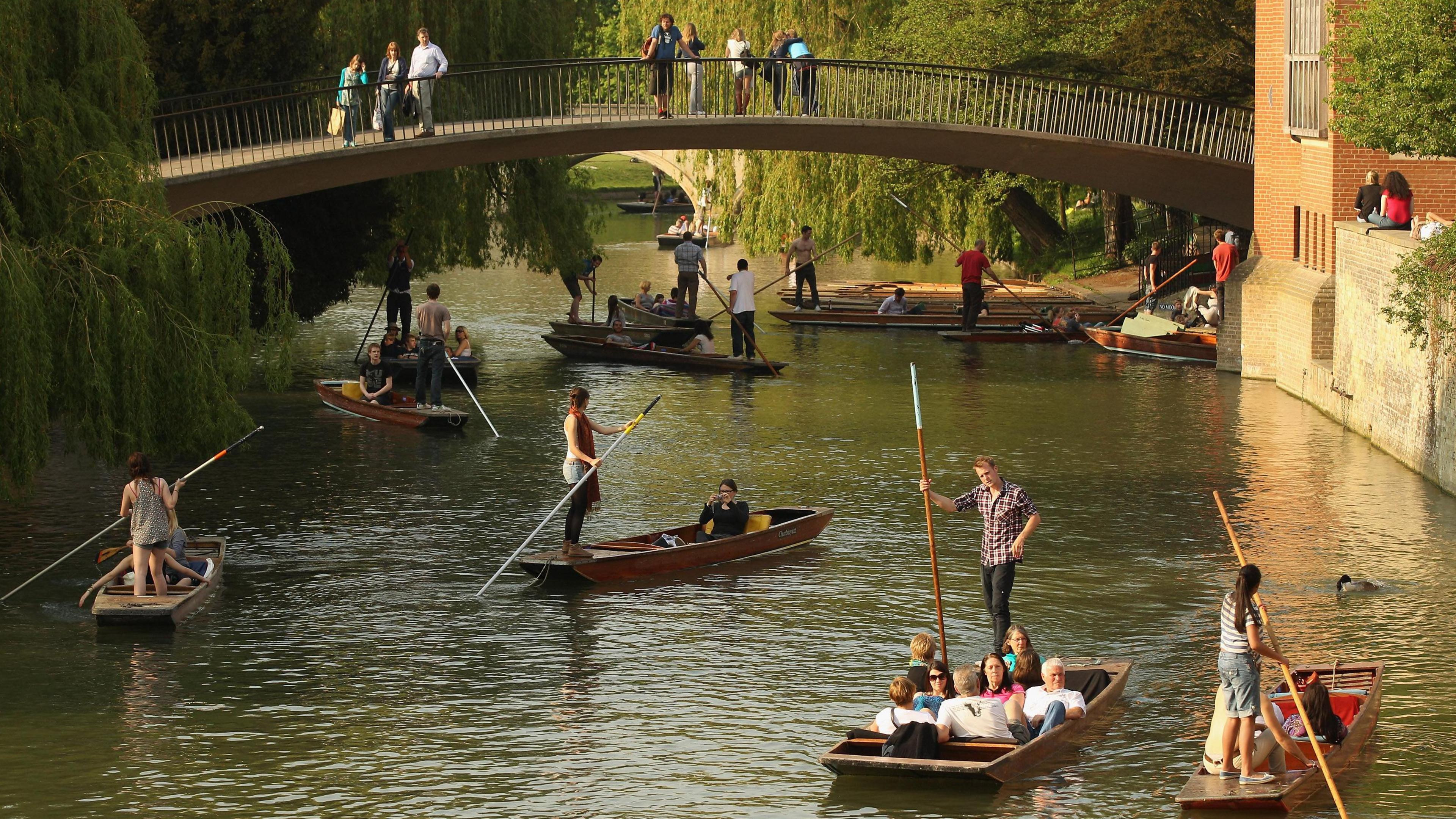Seven or so punts on the River Cam in Cambridge on a sunny day. Some are passing under a curved footbridge which spans the river between weeping willows on the left and buildings on the right.