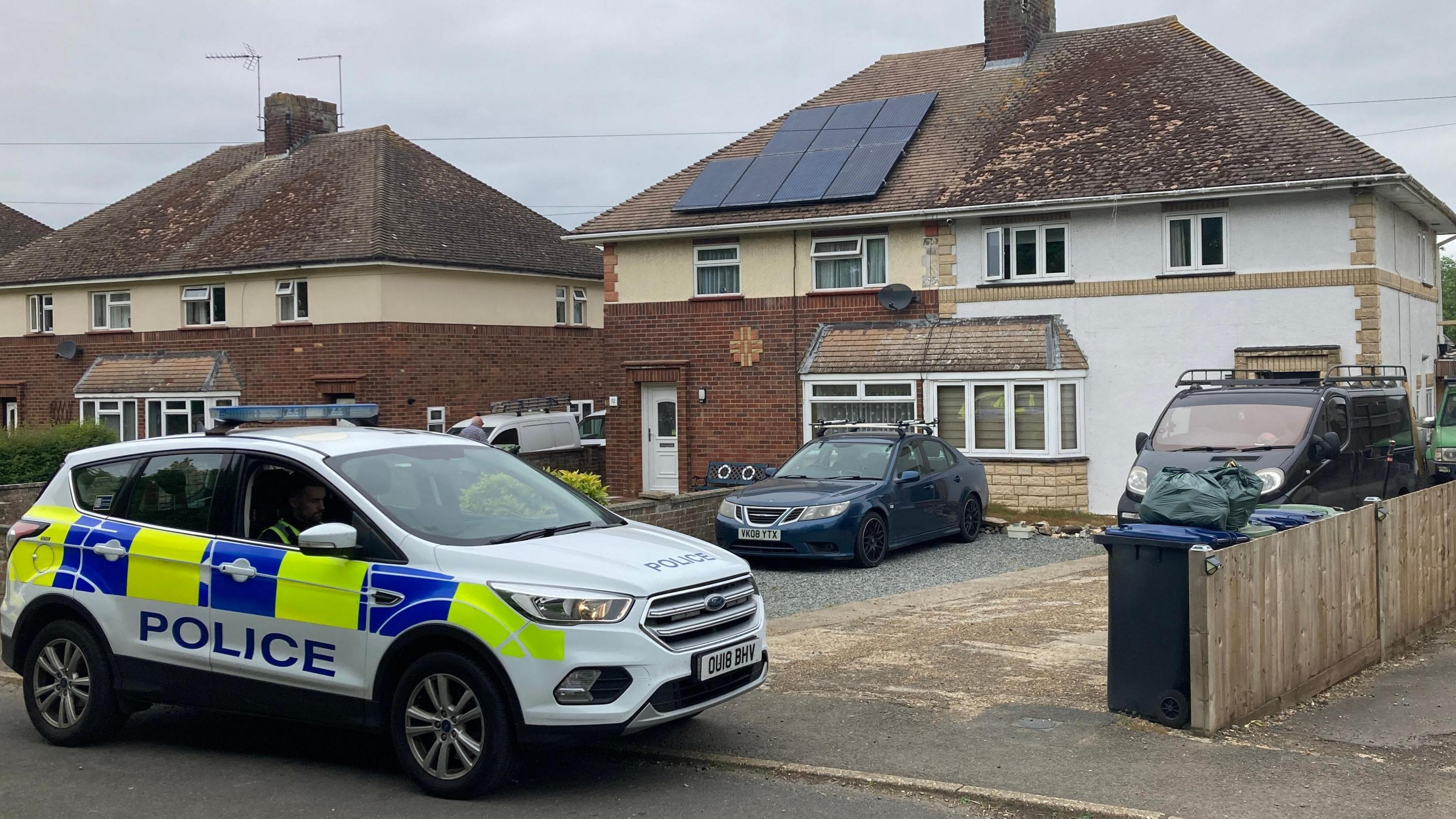 A police car outside a semi-detached white house. It is on the left and a car and a van can be seen on the driveway/paved over garden.