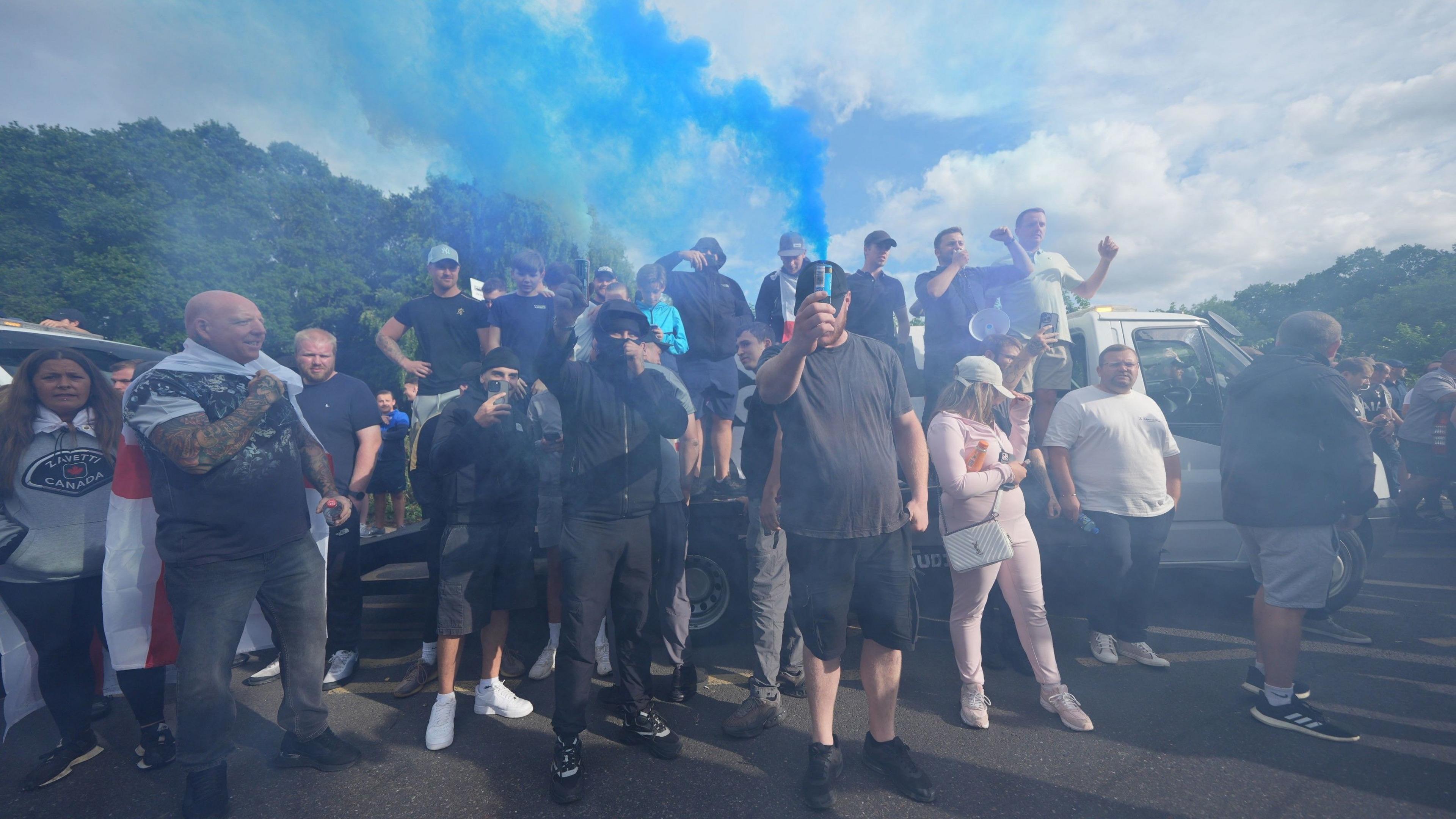 A group of mostly male protesters, some standing on the back of flatbed truck with a white cab. Some are wearing caps or face coverings, while others point their mobile phone cameras towards the photographer. The air is smoky from flares and plumes of blue smoke are rising up from the crowd
