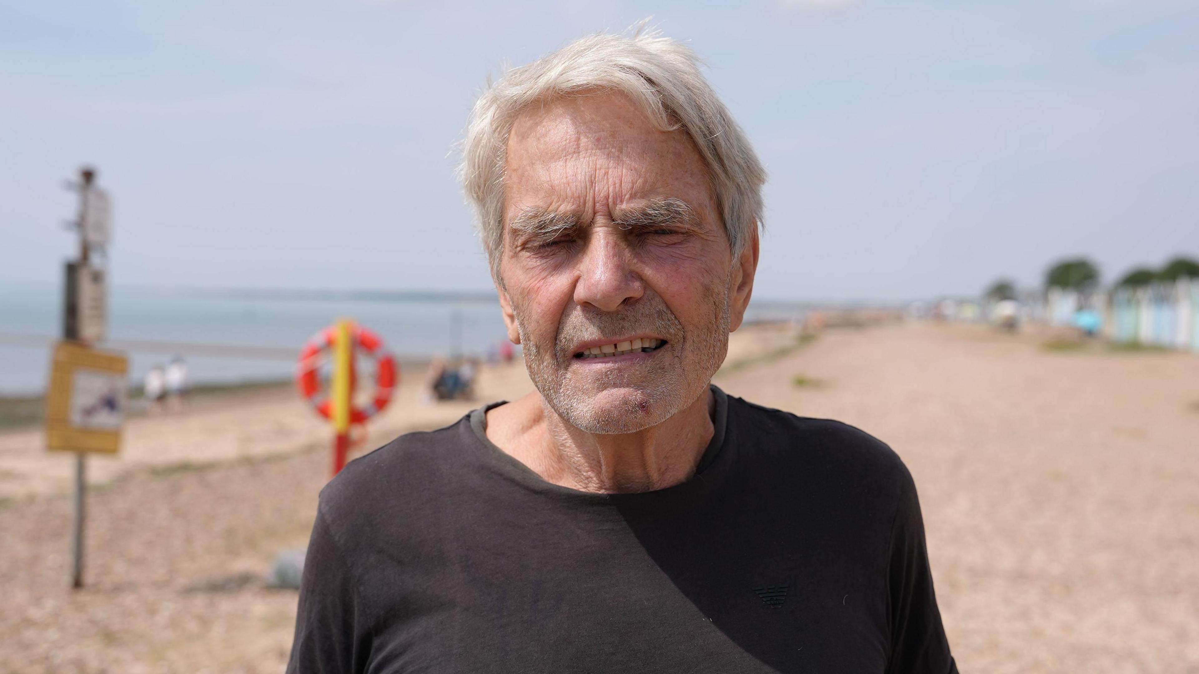 Stephen Vince has grey hair and is wearing a black top. He is standing on the beach.