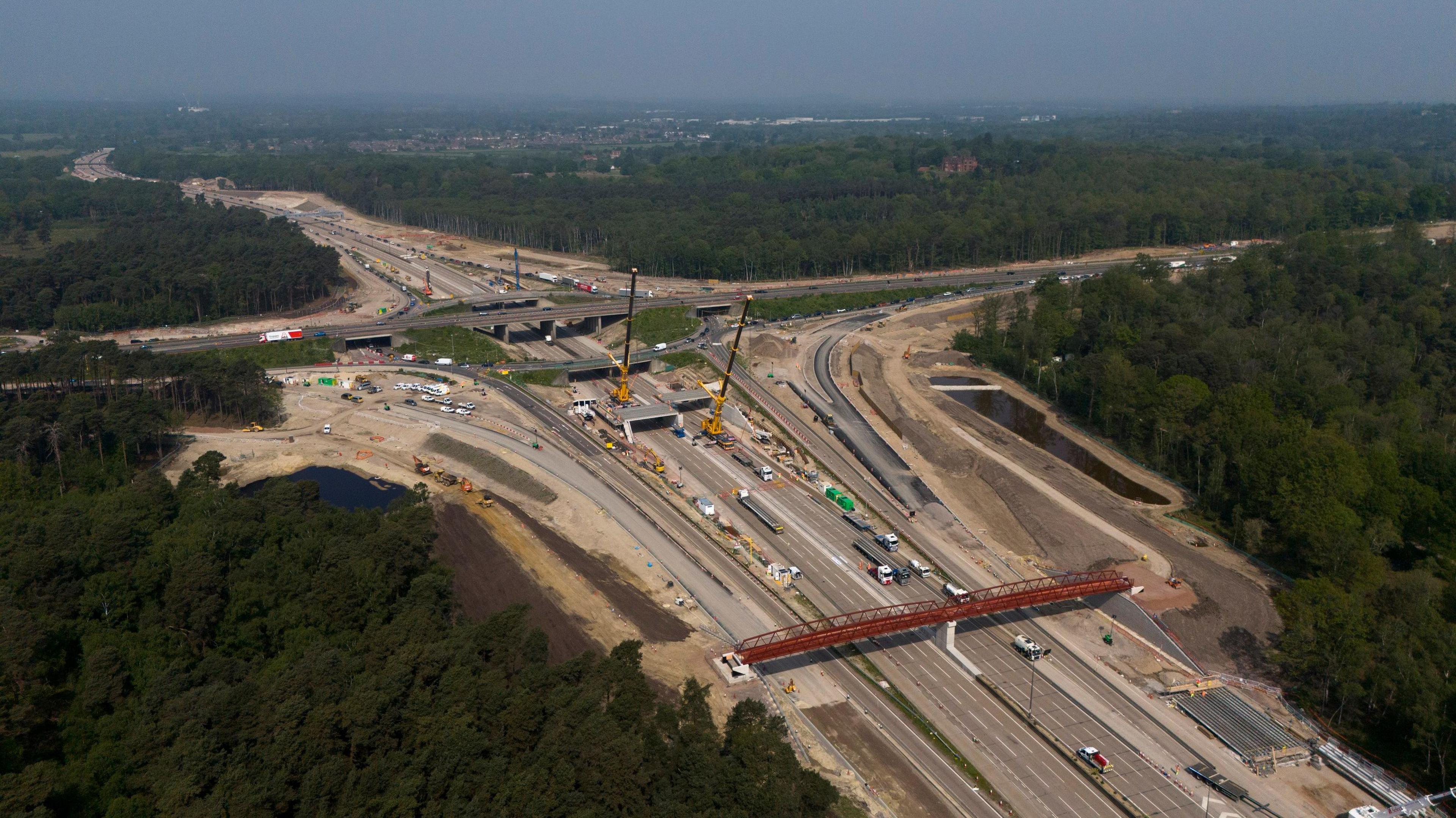 An aerial view showing junction 10 of the A3 and the M25 which has several bridges going over it and with large cranes seen in the middle of the carriageway. There are trees surrounding the large roads which stretch off into the distance.