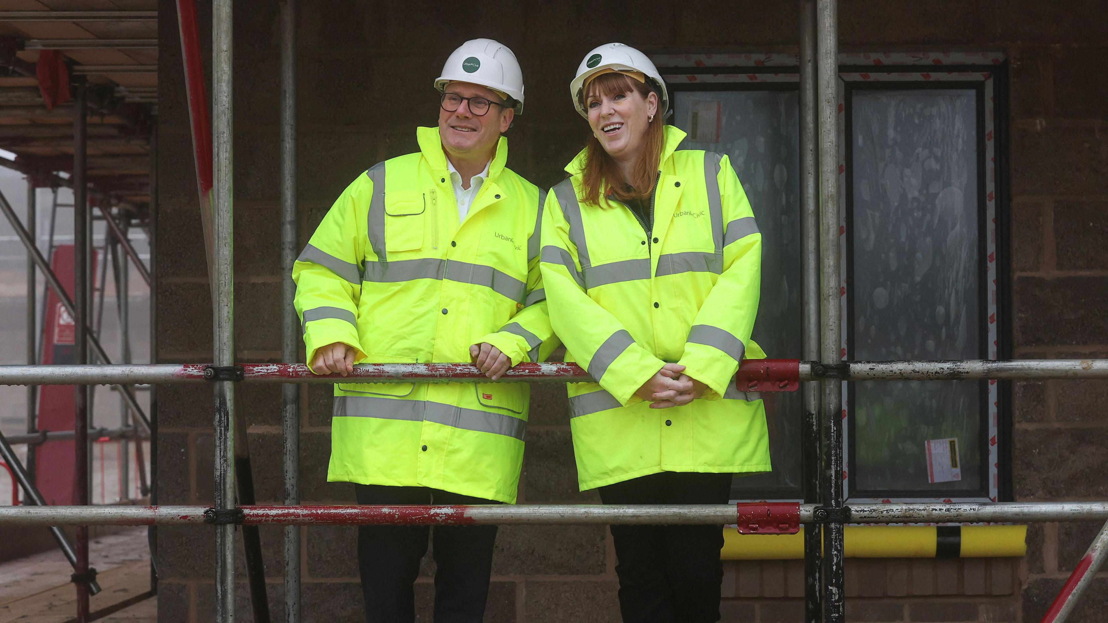 Sir Keir Starmer and Angela Rayner wear high visibility yellow coats and white hard hats. Their hands are placed on a grey and red scaffolding with a partially built building in the background. 