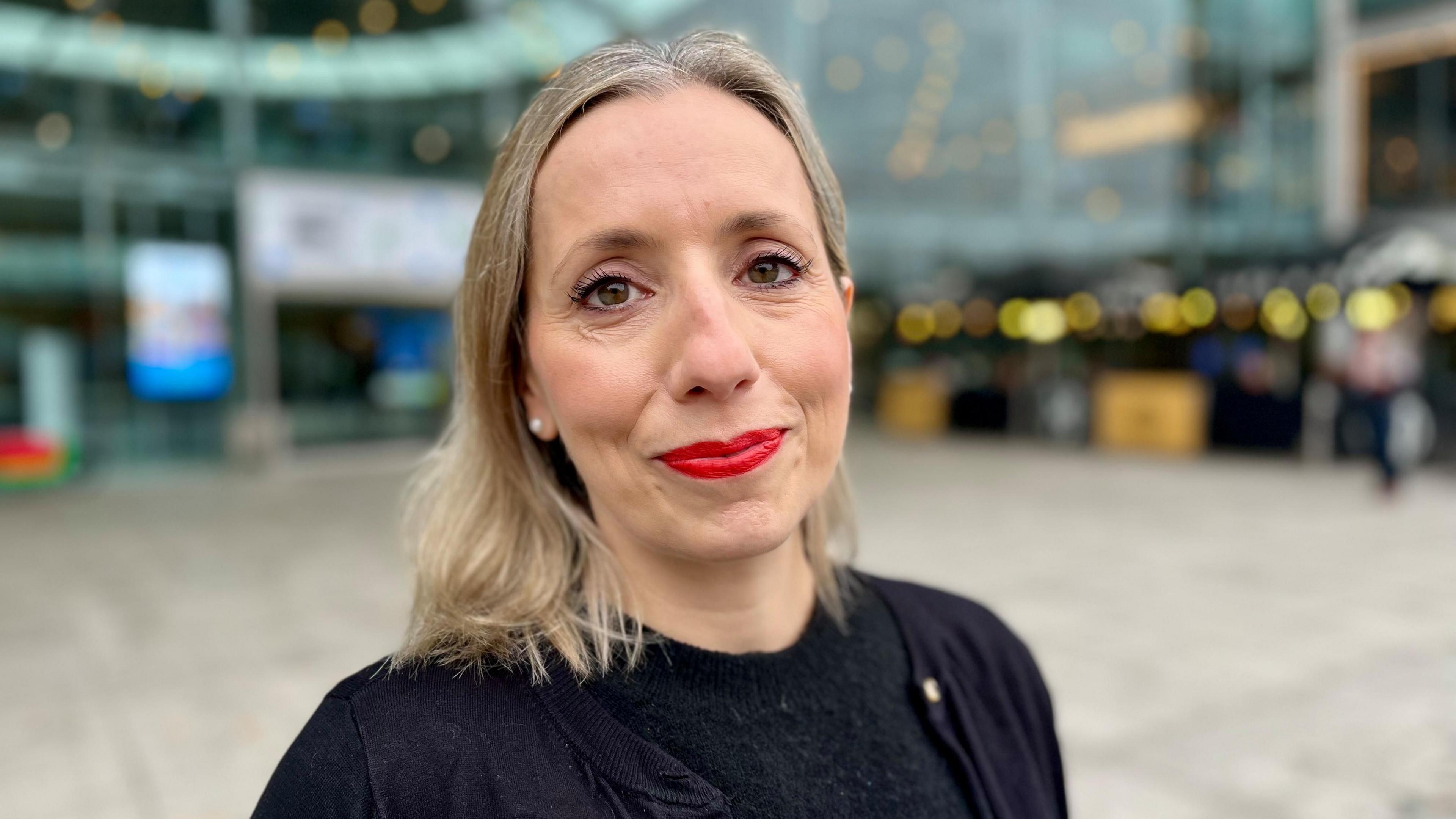 Sarah Taylor with long blond hair and red lipstick smiling at the camera wearing a black top and standing in a large indoor public area with a light-coloured floor