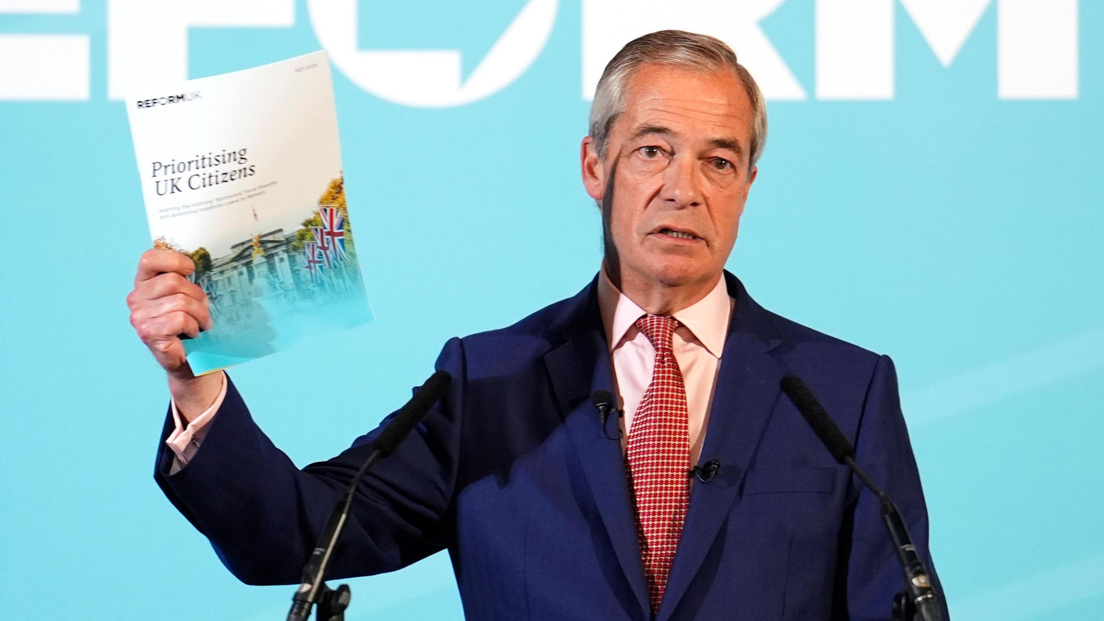 Nigel Farage holds up a piece of paper that reads "Prioritising UK Citizens". He stands in front of two microphones with a pale blue background behind him. The word "reform" can just about be made out above his head, though it is cut off by the border of the photo.