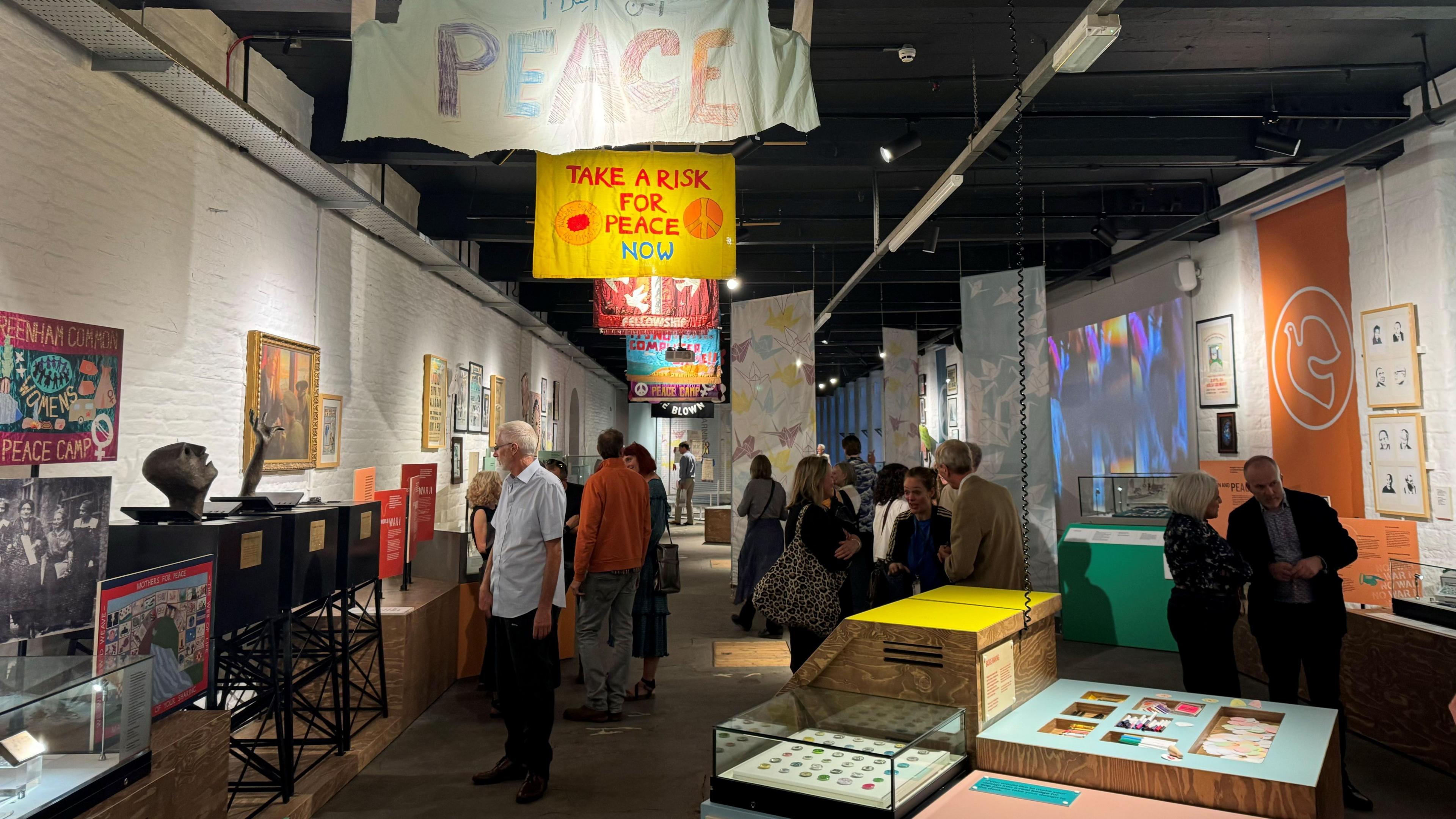 Groups of visitors looking at the Peace Museum's artefacts which are in clear plastic cabinets in the white-washed walled mill room that now houses the collection. Colourful peace banners hang from the ceiling.  