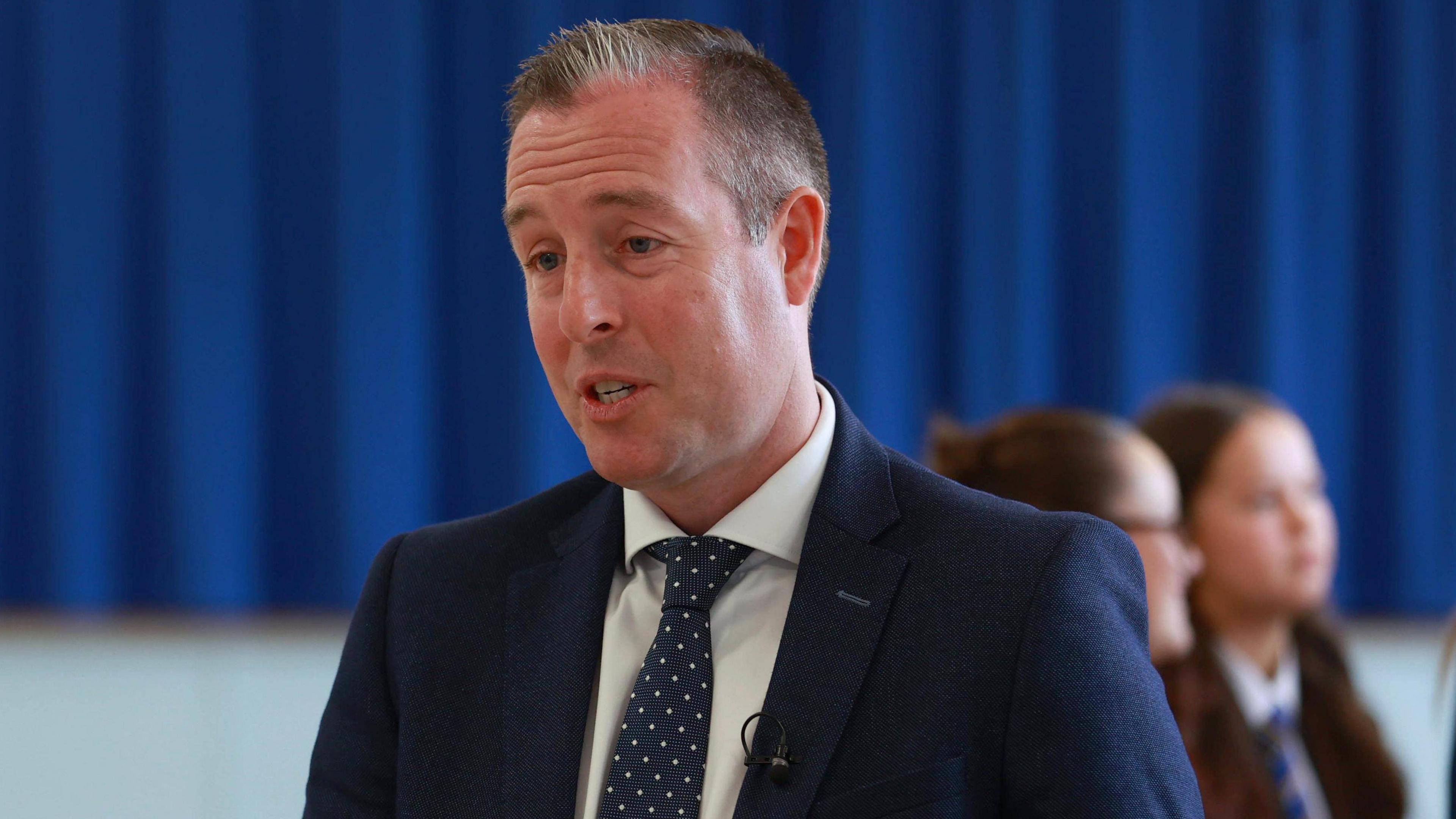 A man in a navy suit, white shirt and blue tie has short grey hair. Behind him, out of focus, are two young girls with brown hair.