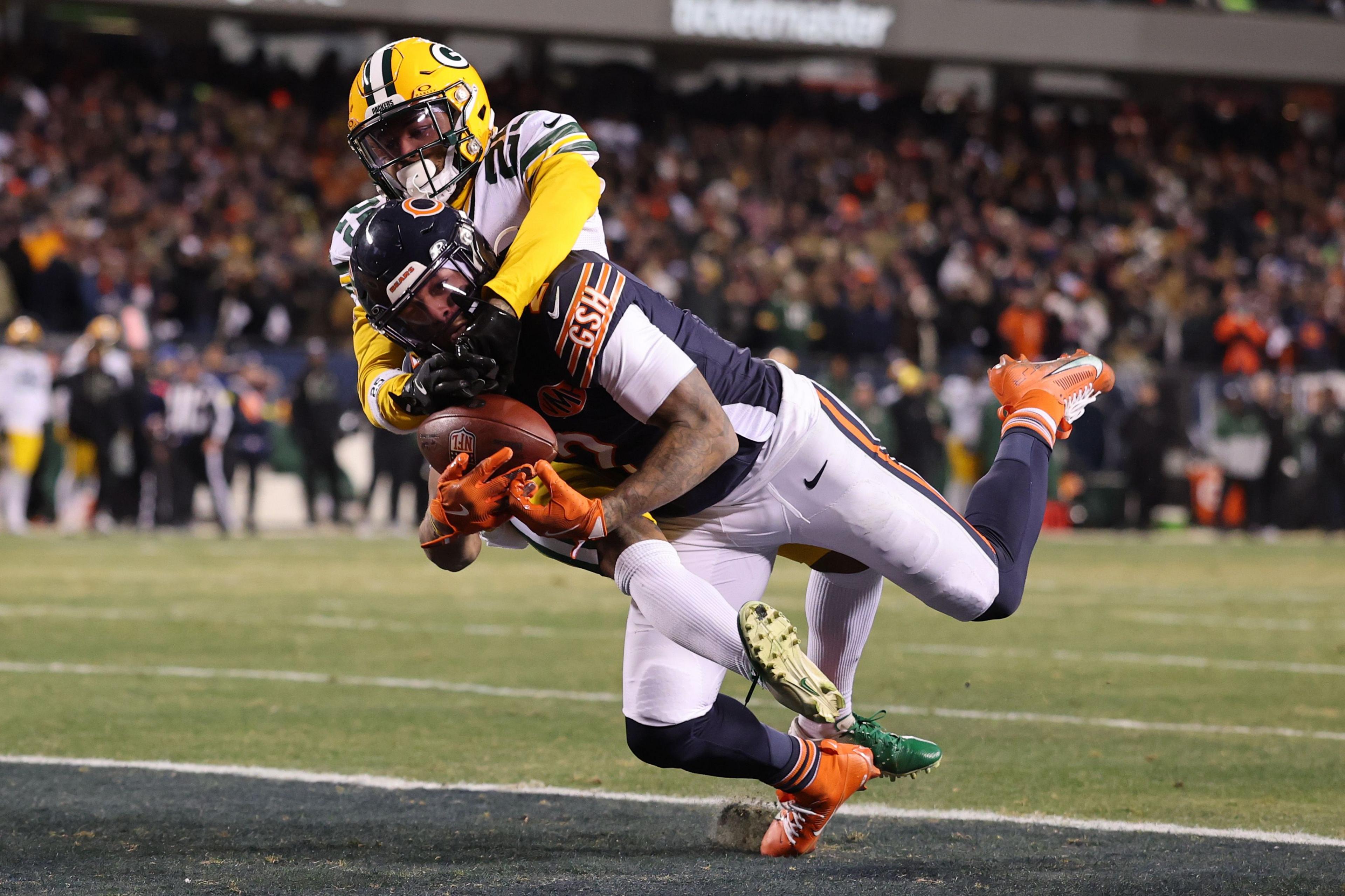 An American football player in a dark navy uniform is diving forward while holding the ball tightly, being tackled by an opposing player in a green and yellow uniform. The action takes place near the end zone on a grassy field, with a blurred crowd in the background.