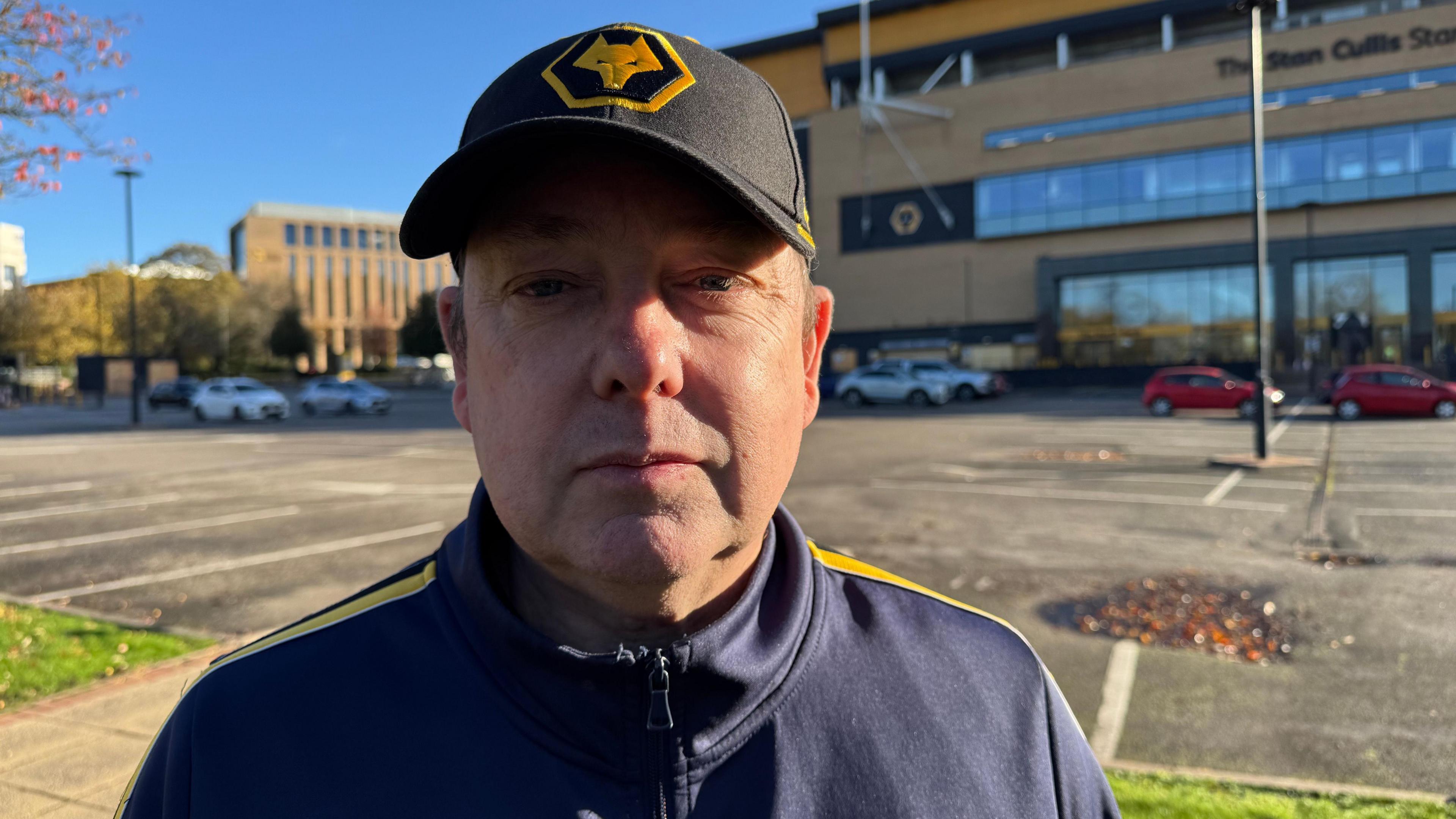Dave Milton standing outside The Molineux Stadium. It is a huge building and we can see the Wolves logo. He is a middle aged white man. He is wearing a Wolves hat and a blue and golden jacket. 