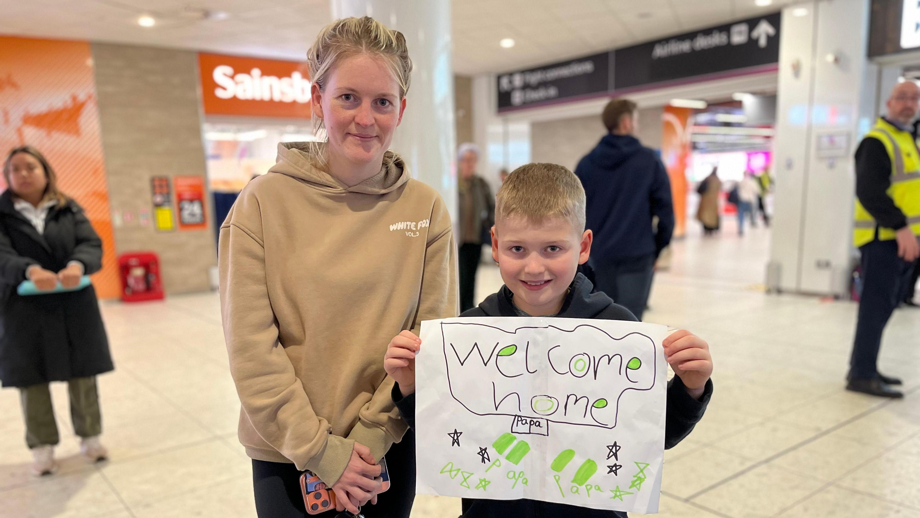Një nënë dhe bir presin një anëtar të familjes në Aeroportin e Edinburgut. Ata mbajnë lart një tabelë që thotë "Mirë se erdhe në shtëpi, baba".