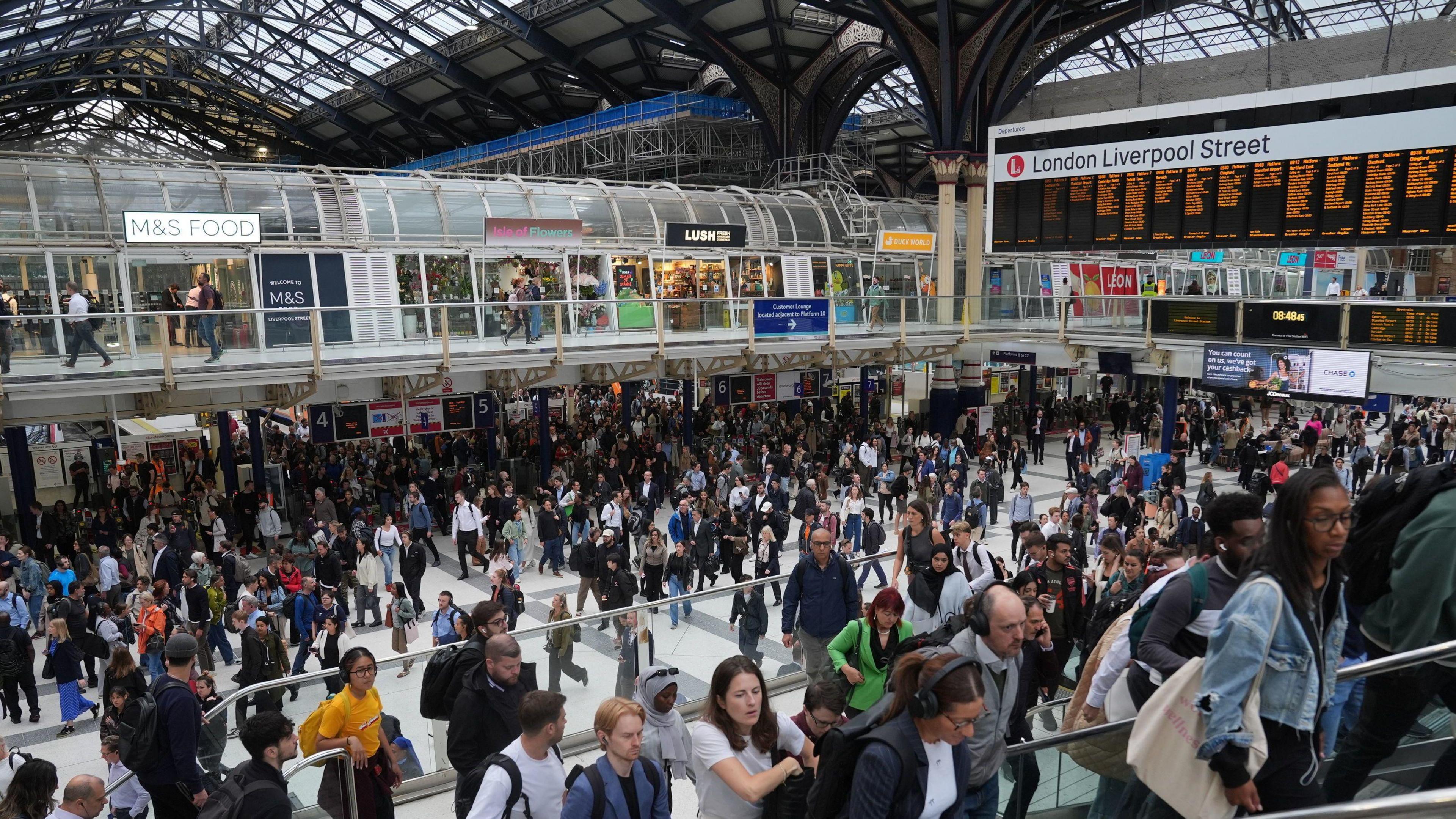 Liverpool Street Station concourse. You can see shops, hundreds of people on the concourse and escalator and departure boards.