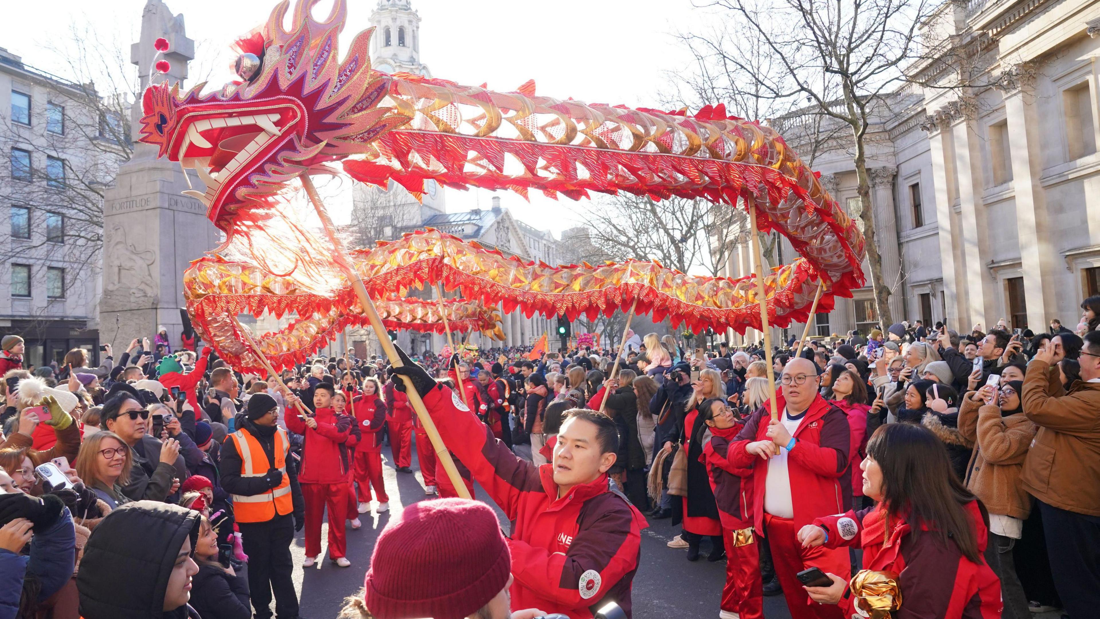 A large crowd watches a Lunar New Year parade featuring performers carrying a long, golden-red dragon puppet. The event takes place in a central London street.