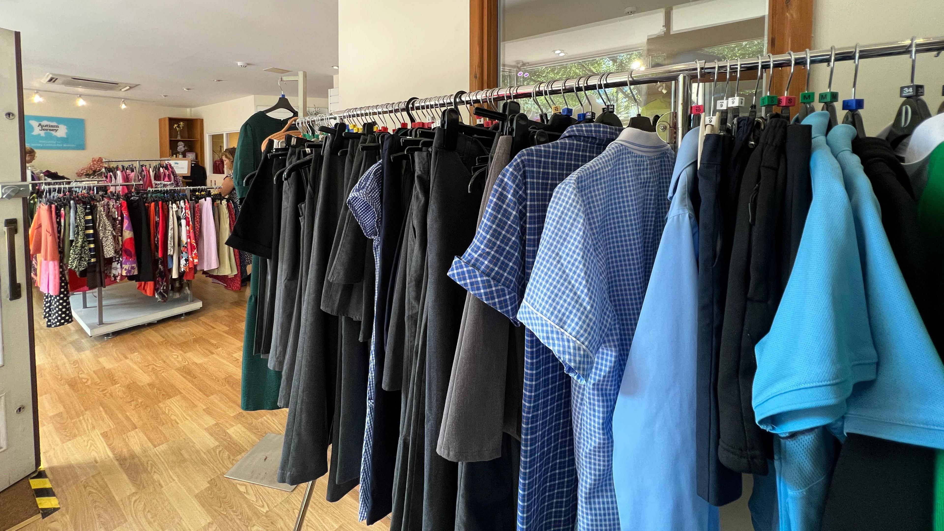 Interior of a clothing store with wooden flooring, featuring racks of shirts and jackets in the foreground, including blue checkered and solid-colored tops. Additional racks with dresses and other garments are visible in the background. The store is well-lit with natural light.