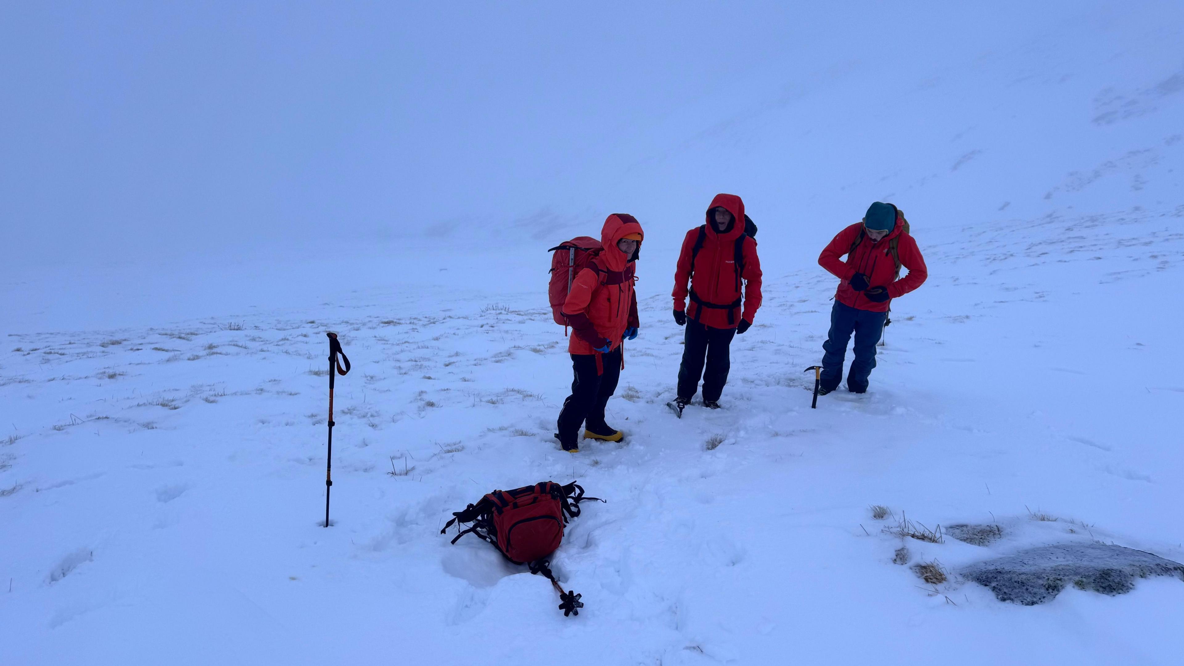 A group of three walkers stand in snow. They are wearing winter climbing clothing and two of them have their hoods up. There is a rucksack and a pair of walking poles on the snow-covered ground. 