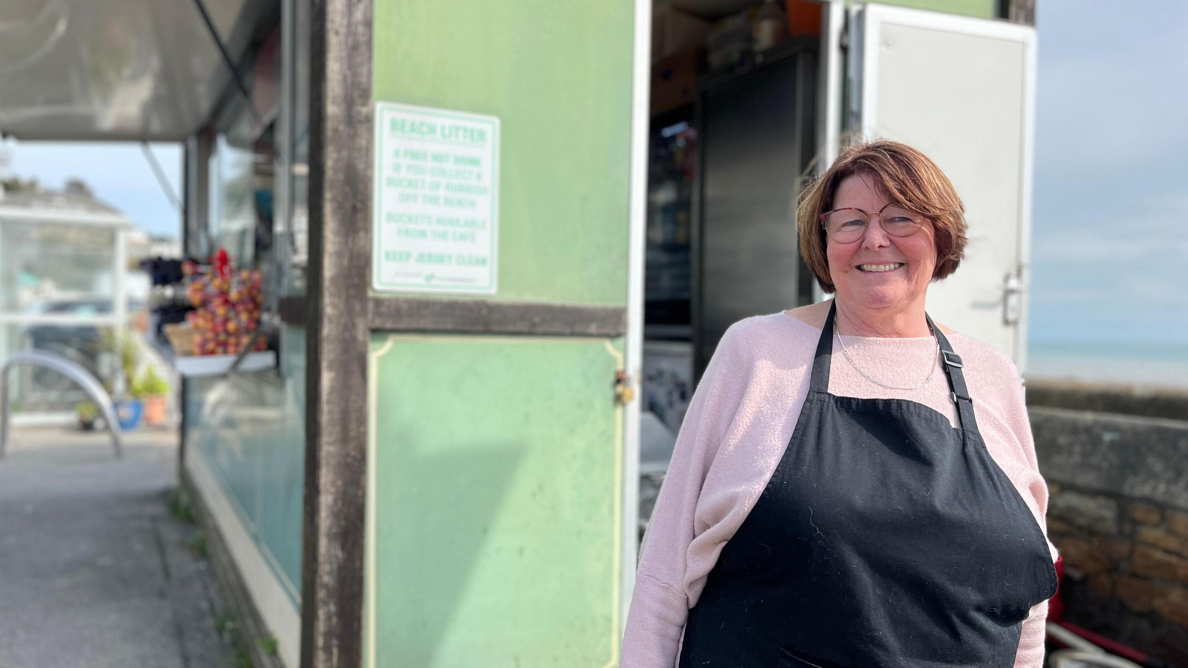 Rhona Hewit, a woman in a light pink sweater and black apron, stands outside a small green food kiosk with an open door. A sign on the wall warns against beach littering and encourages proper bin use to keep Jersey clean. The surrounding area includes some plants and an outdoor setting.