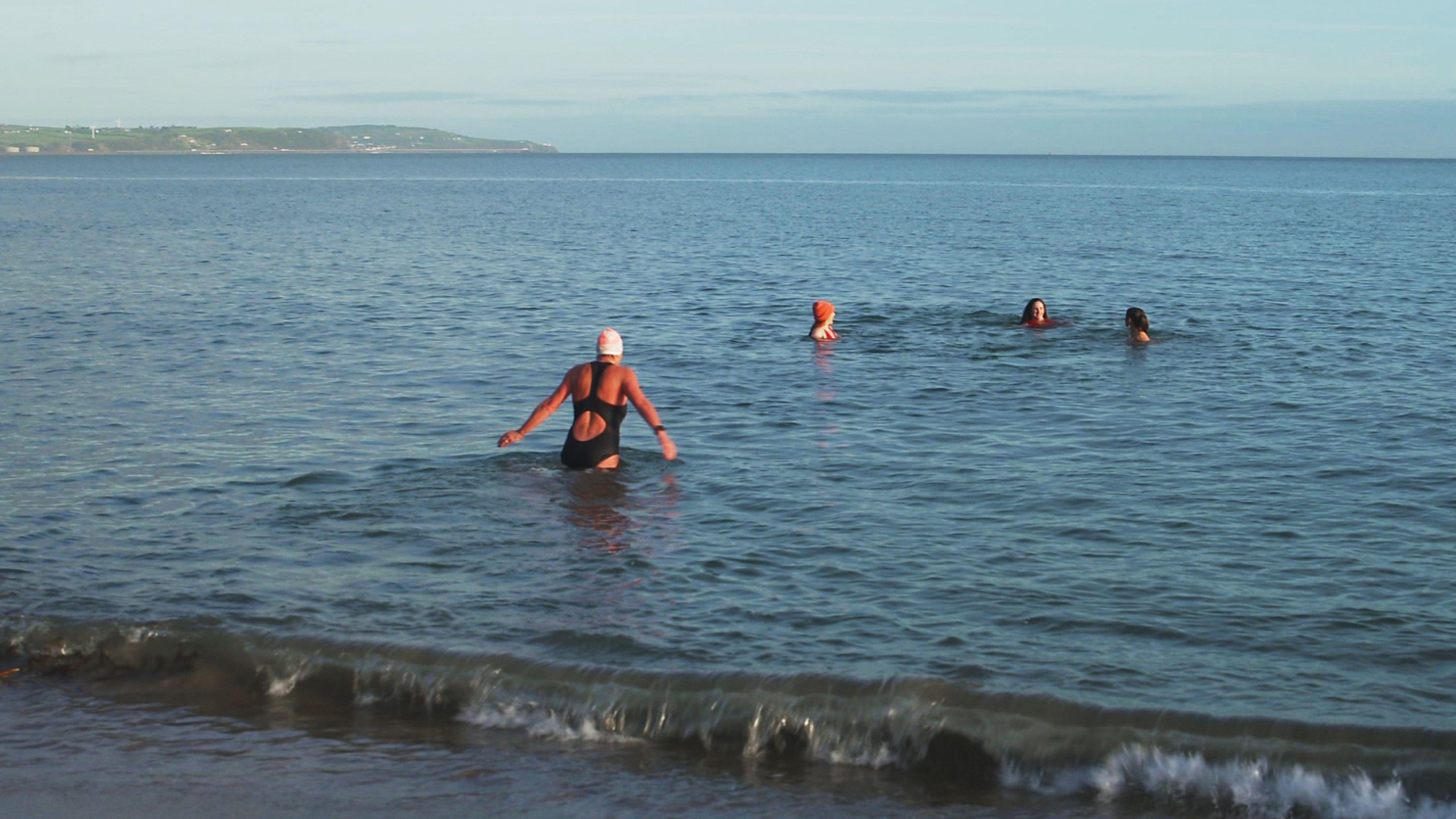 A woman in a black swim suit and white swim cap wades into the sea as a rolling wave crashes on the shore. Three other women are already mostly submerged in the water with their heads visible. Two of the women on the right have dark hair, the other is wearing an orange wooly hat and a red swim suit. Some land is visible to the left in the background showing another beach and some houses and fields. 