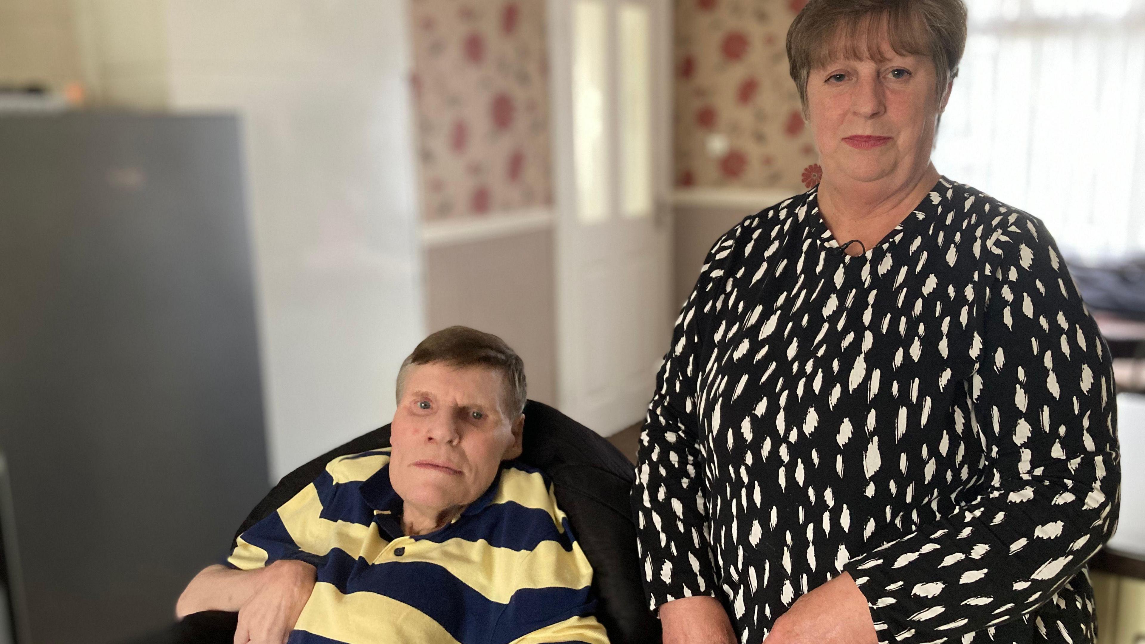 A man with a yellow and blue jumper sat next to a woman with short light brown hair wearing a black dress with white spots who is standing up.