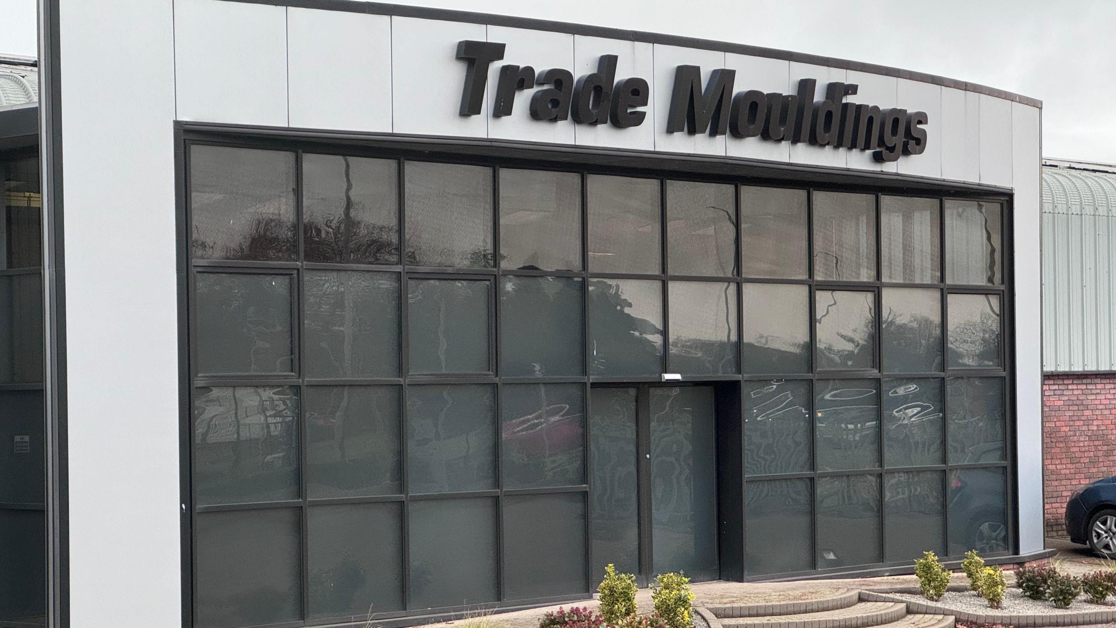 The front of the Trade Mouldings factory in Cookstown.  The cladded building has a large wall of blacked-out windows at its entrance and a large black sign showing the firm's name. 