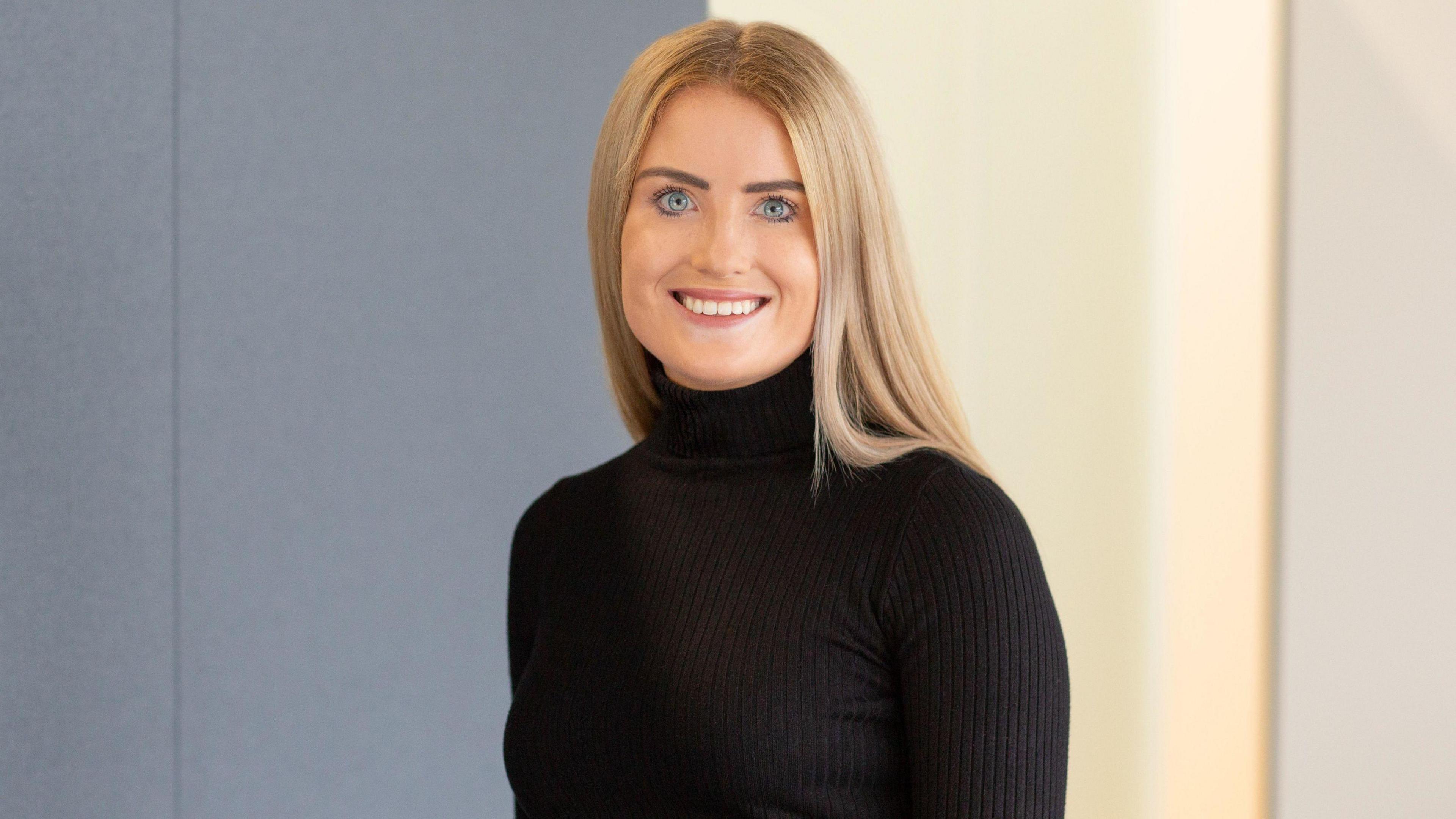 A caucasian girl in her 20s with long blonde hair and blue eyes smiles at the camera in a professional head-shot whilst wearing a black turtle-neck sweater.