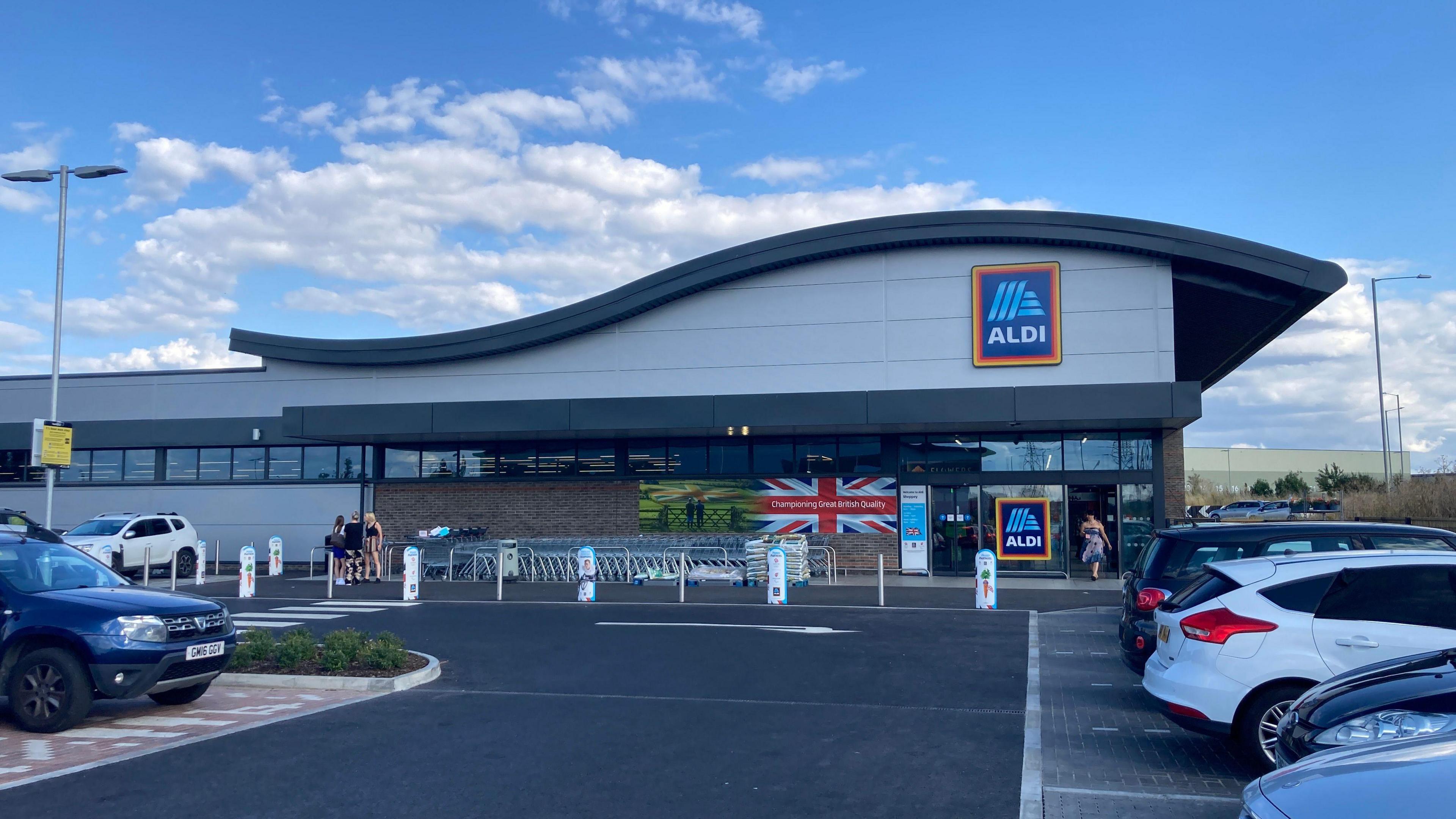 An Aldi store. The black roof is shaped like a wave. The res of the building is grey. In the foreground is black tarmac of the car park. There are four cars parked on the right, one on the left. The sky is blue, a few clouds scattered in the background.