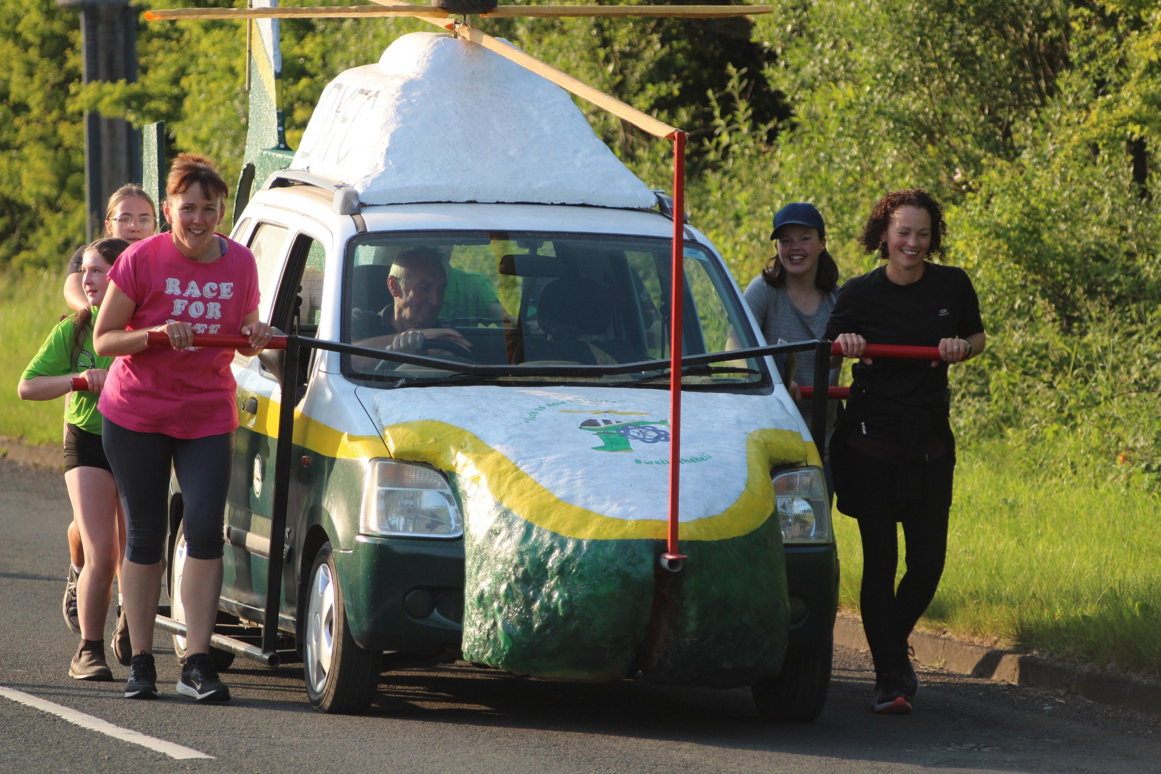 Six people are walking beside the replica helicopter, pulling it along using metal rods attached to the car. They are smiling as they pull it along the rural road.