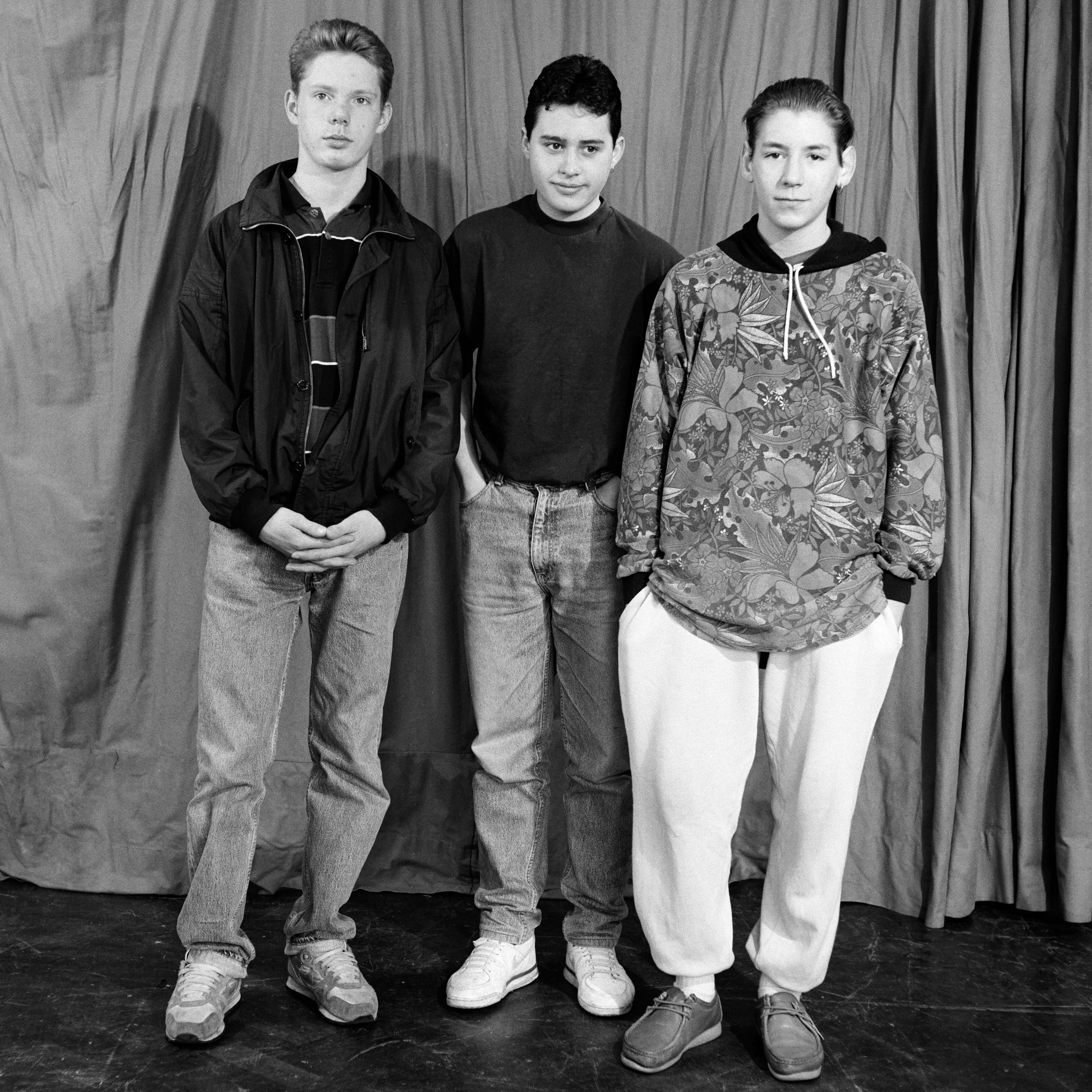 Boys from Tulse Hill School, 1990. Three young men stand in front of a curtain. Two wear jeans, one wears tracksuit bottoms. They wear jumpers and jackets. Black and white photo.