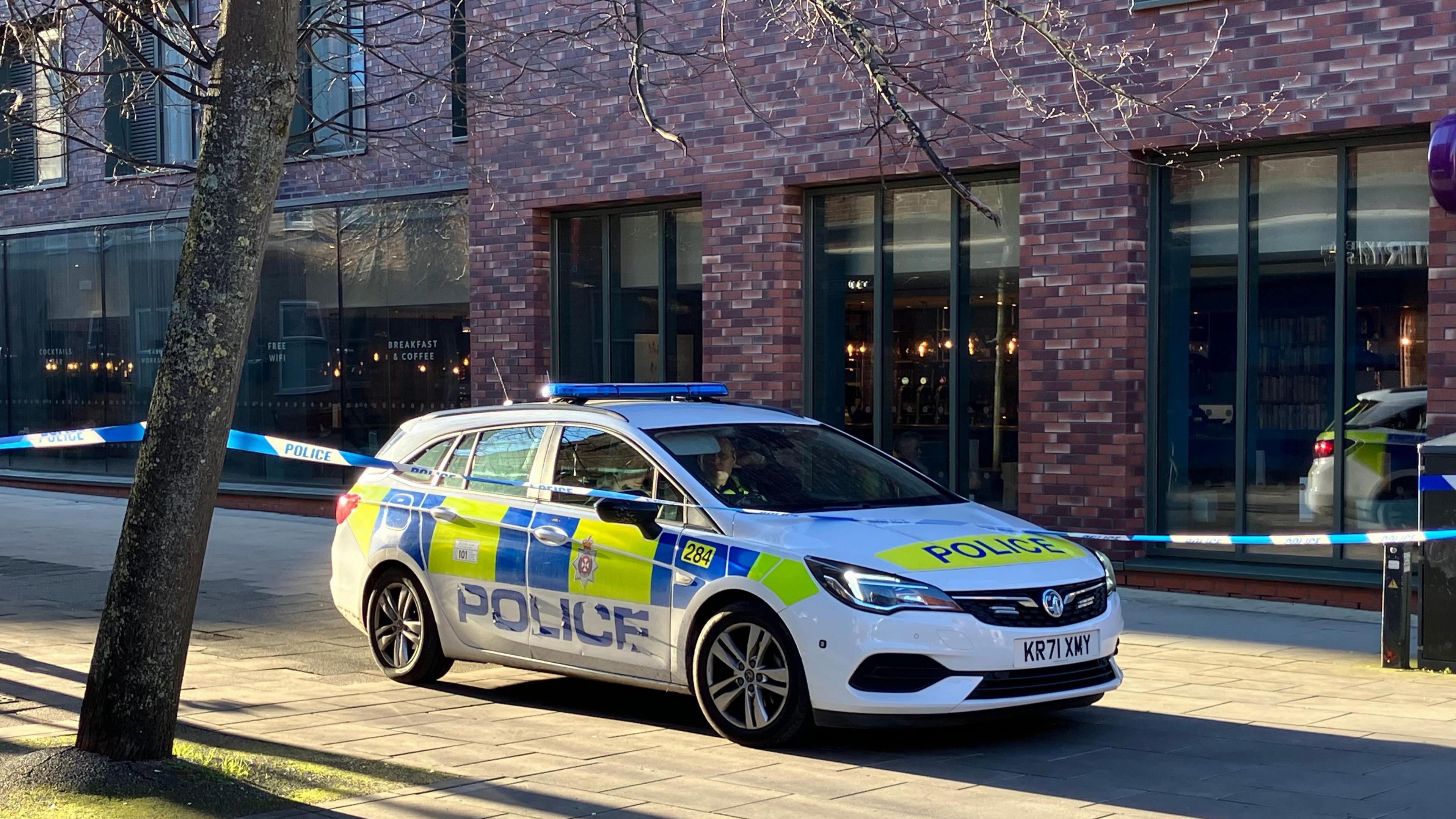 A police car behind police tape on a paved street, in front of a modern brick building with large windows and tree to the left.