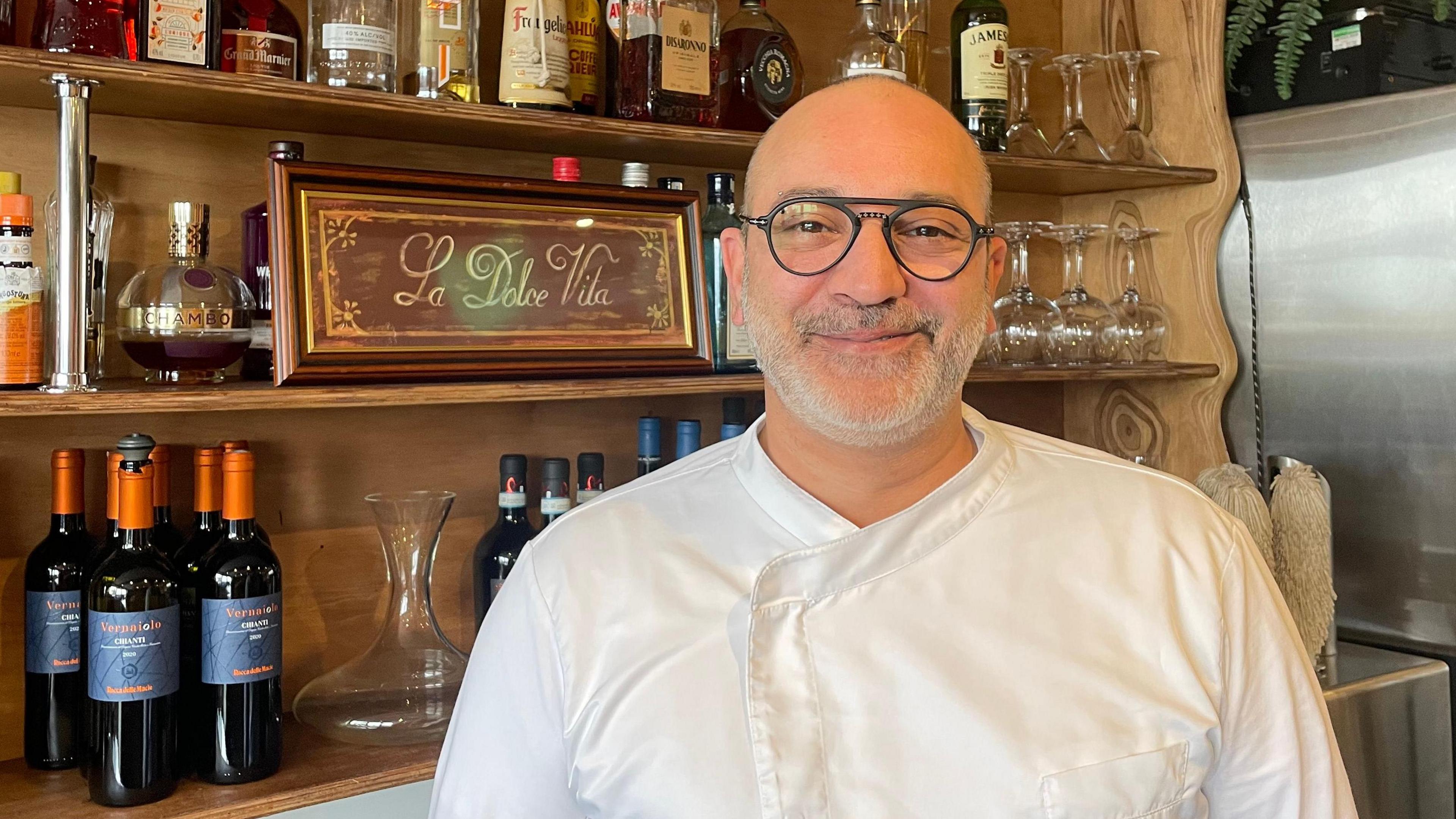 Giuseppe Catalano, a chef at Italian restaurant La Dolce Vita, smiling at the camera. He is wearing a white chef's outfit, and standing in front a cabinet with a sign saying La Dolce Vita, and a variety of bottles, glasses and other drinks containers