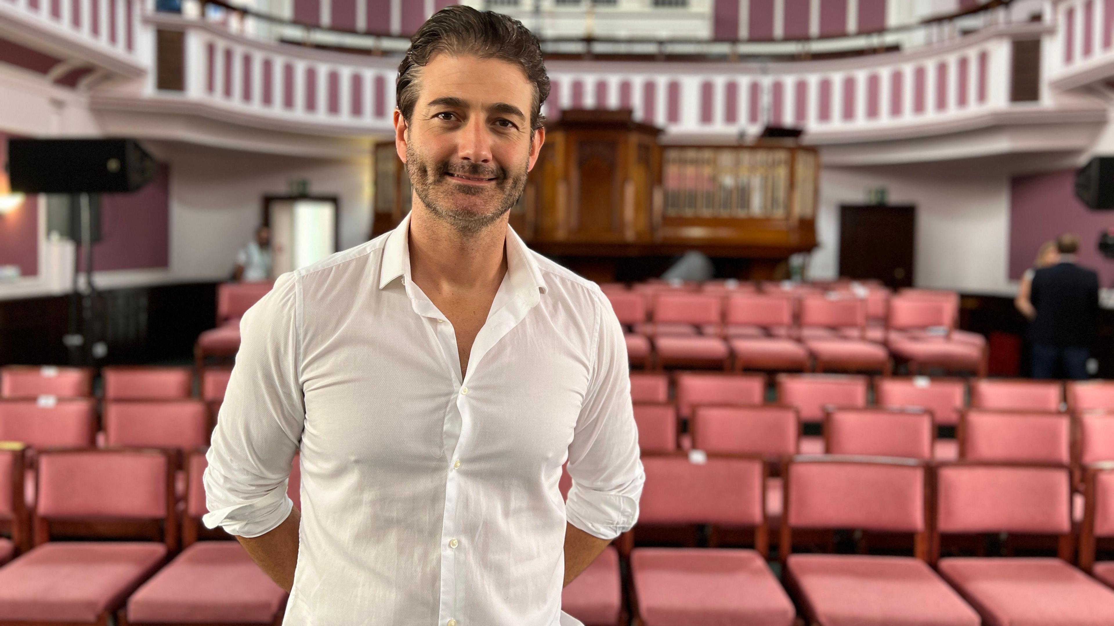 Oliver Tompsett is smiling as he stands in front of rows of seating in a performance space. He has short brown hair and is wearing a white shirt. 