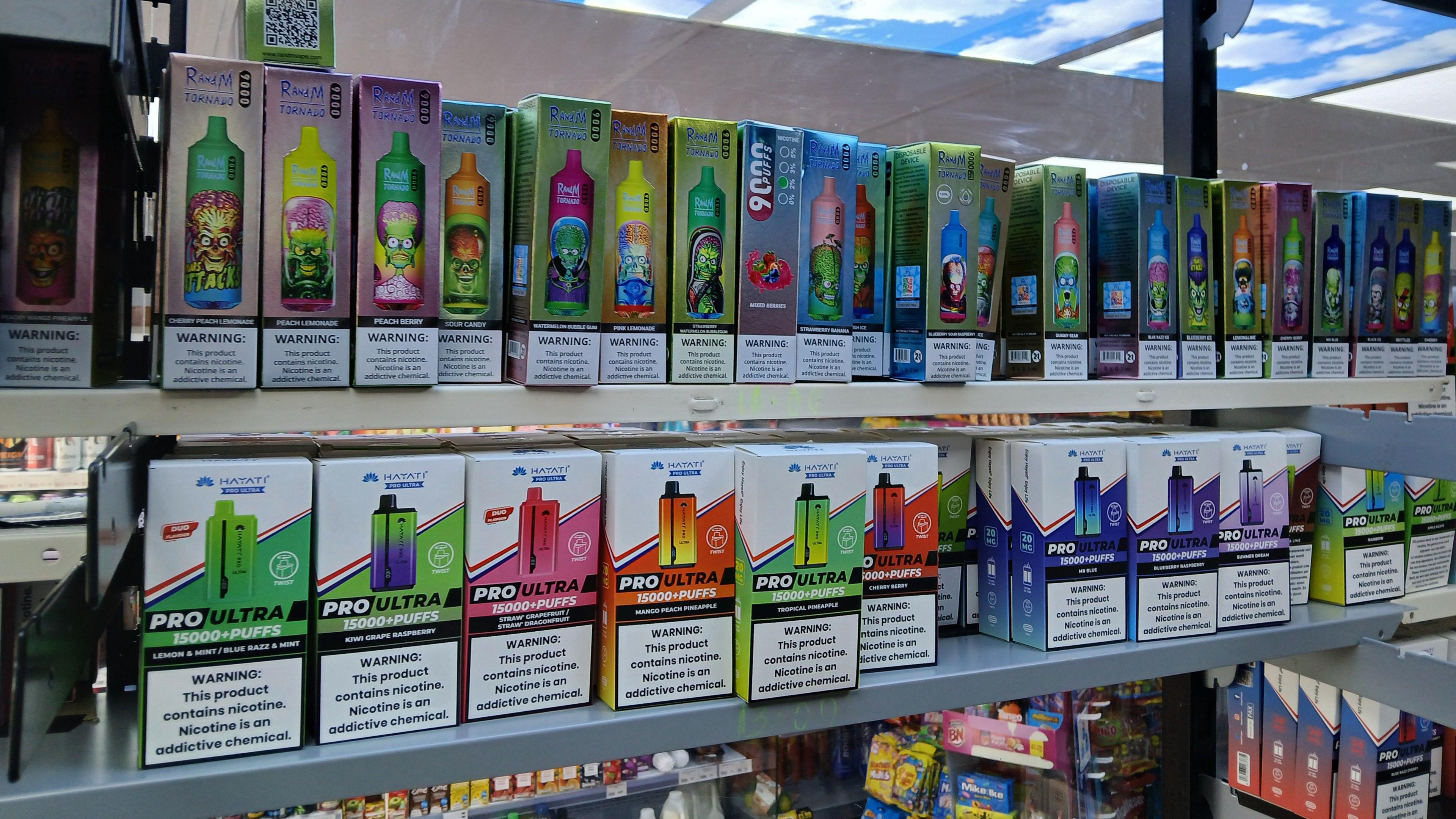 A shelving unit in a store packed with vapes and cigarettes. They range inc colours and are stacked across grey, metal shelves.