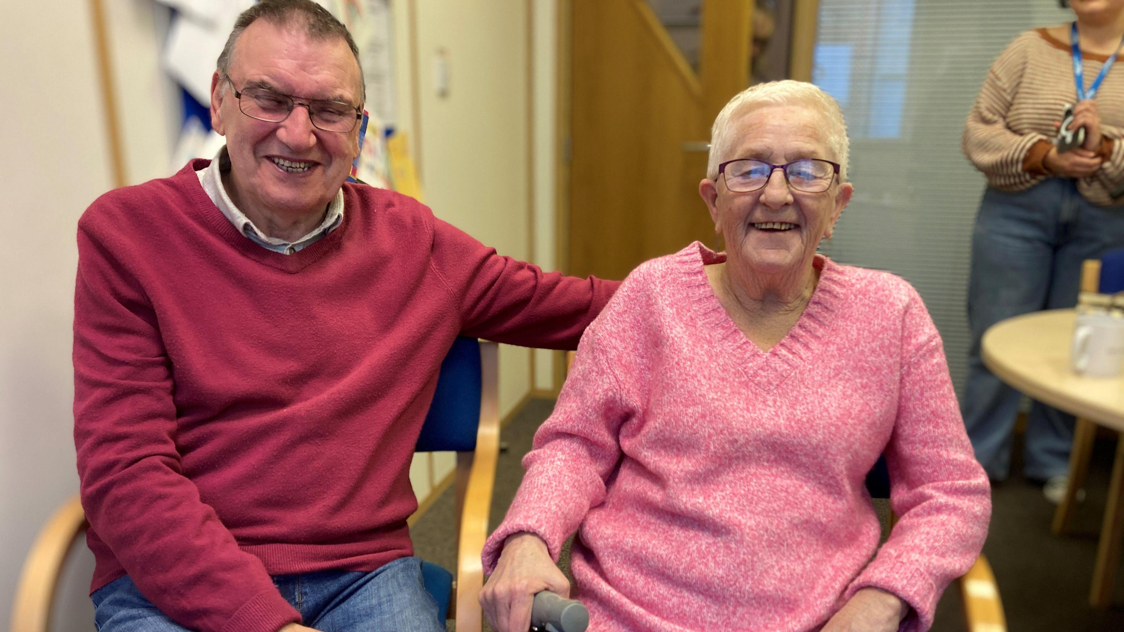 Eddie and Maggie sit side by side in an office. They are both smiling broadly at the camera. He is wearing a red jumper and she is wearing a pink jumper.