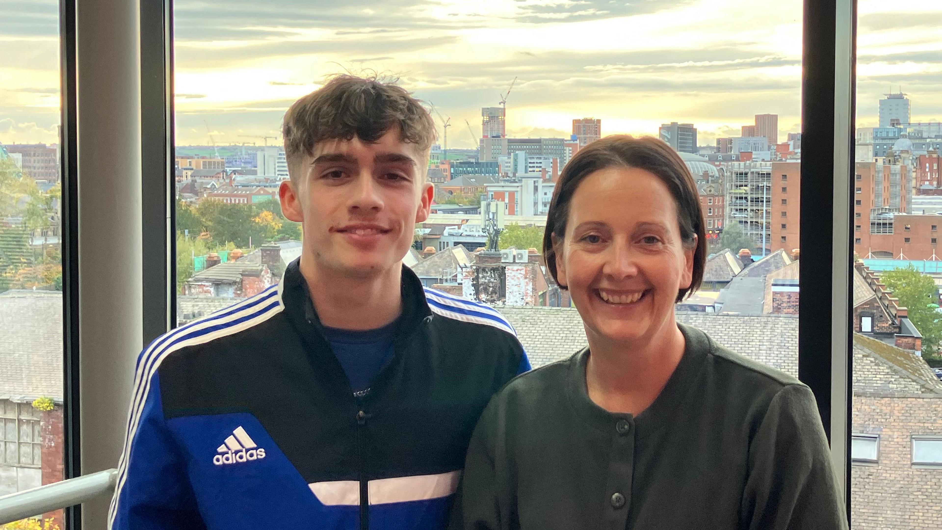 Joe and Tracy are standing indoors in front of large floor-to-ceiling windows with a cityscape visible in the background. The view outside shows a mix of modern and older buildings, rooftops, and construction cranes under a partly cloudy sky with warm light from the setting or rising sun. Joe is wearing a dark jacket with blue and white stripes and an Adidas logo, and Tracy is dressed in a plain dark top. They are positioned close together, with the urban skyline stretching across the horizon behind them.