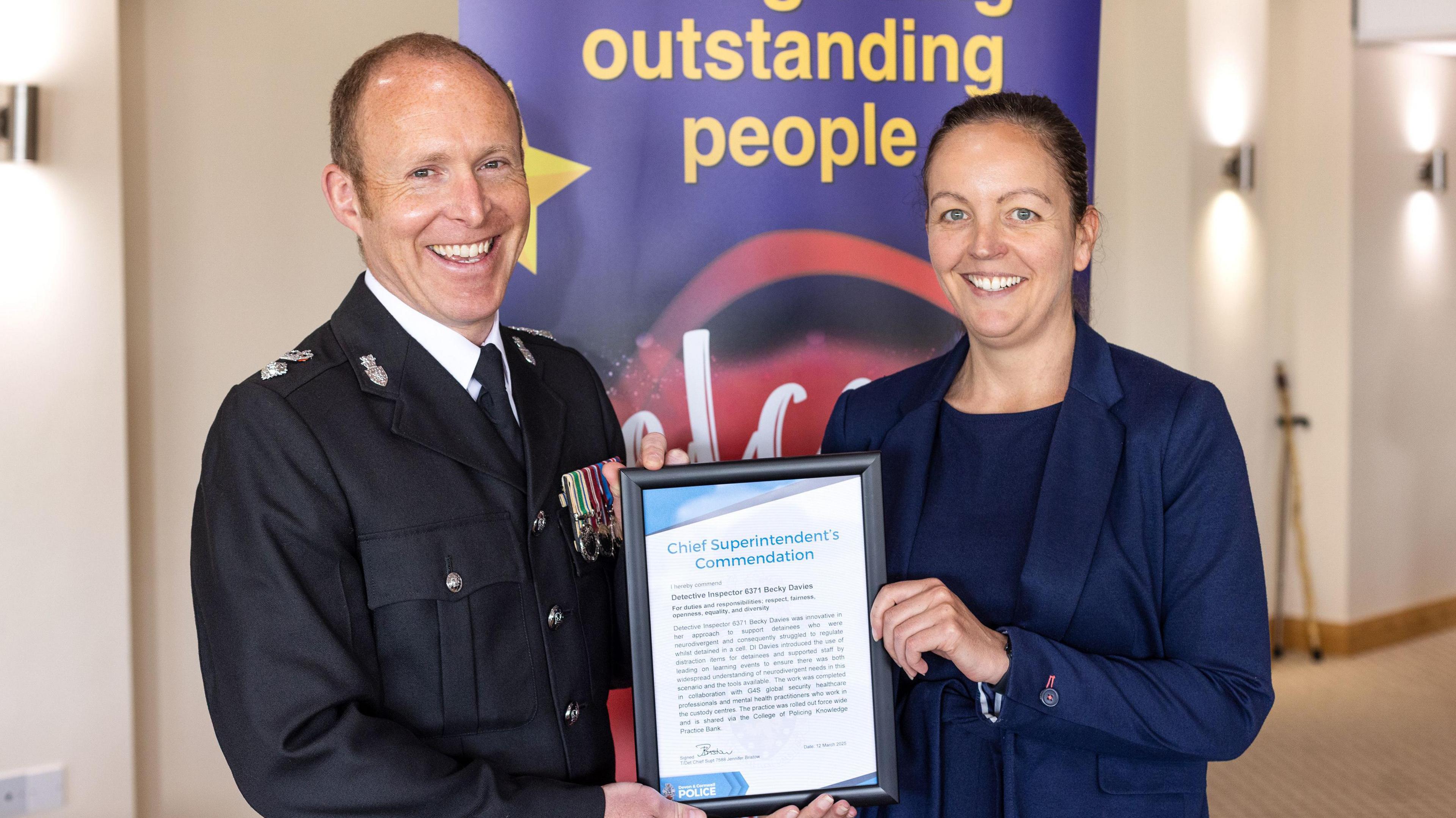 Chief Supt Roy Linden in police uniform, South Devon Police Commander, (left) giving a certificate to Det Ch Insp Becky Davies wearing a navy blue shirt and blazer. Both are smiling. Behind them is a sign which says "outstanding people". The wall behind it is cream. 