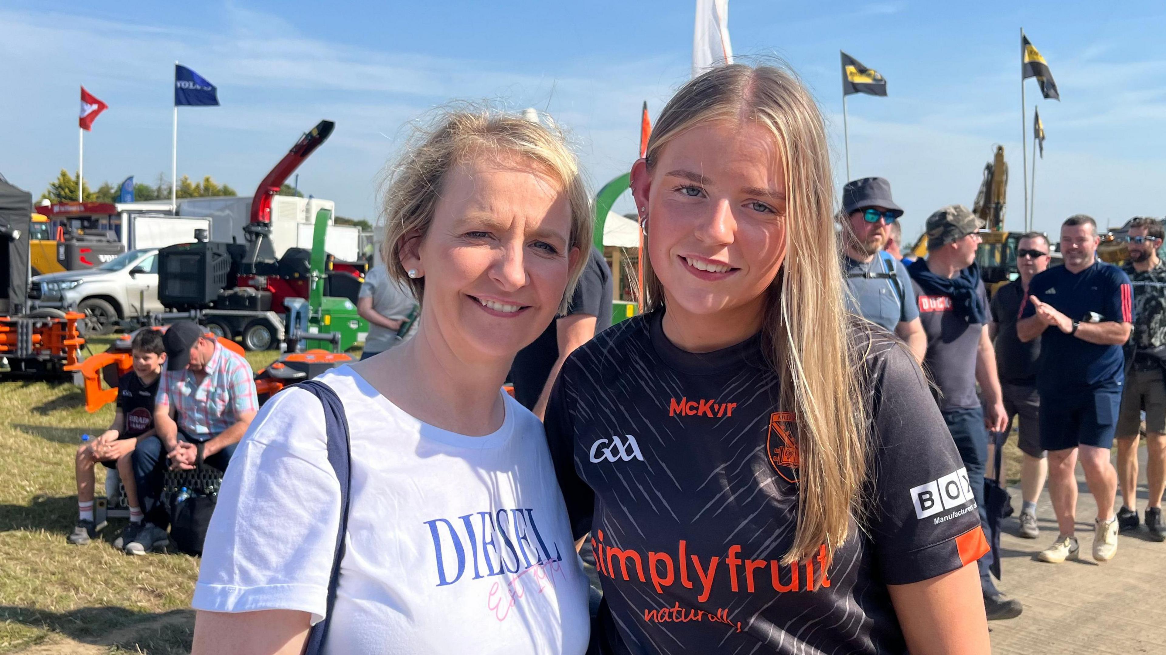 Caroline, with blonde hair, is wearing a white T-Shirt with "diesel" written on the front. Síofra, with blonde hair, is wearing a black GAA top with orange detail.