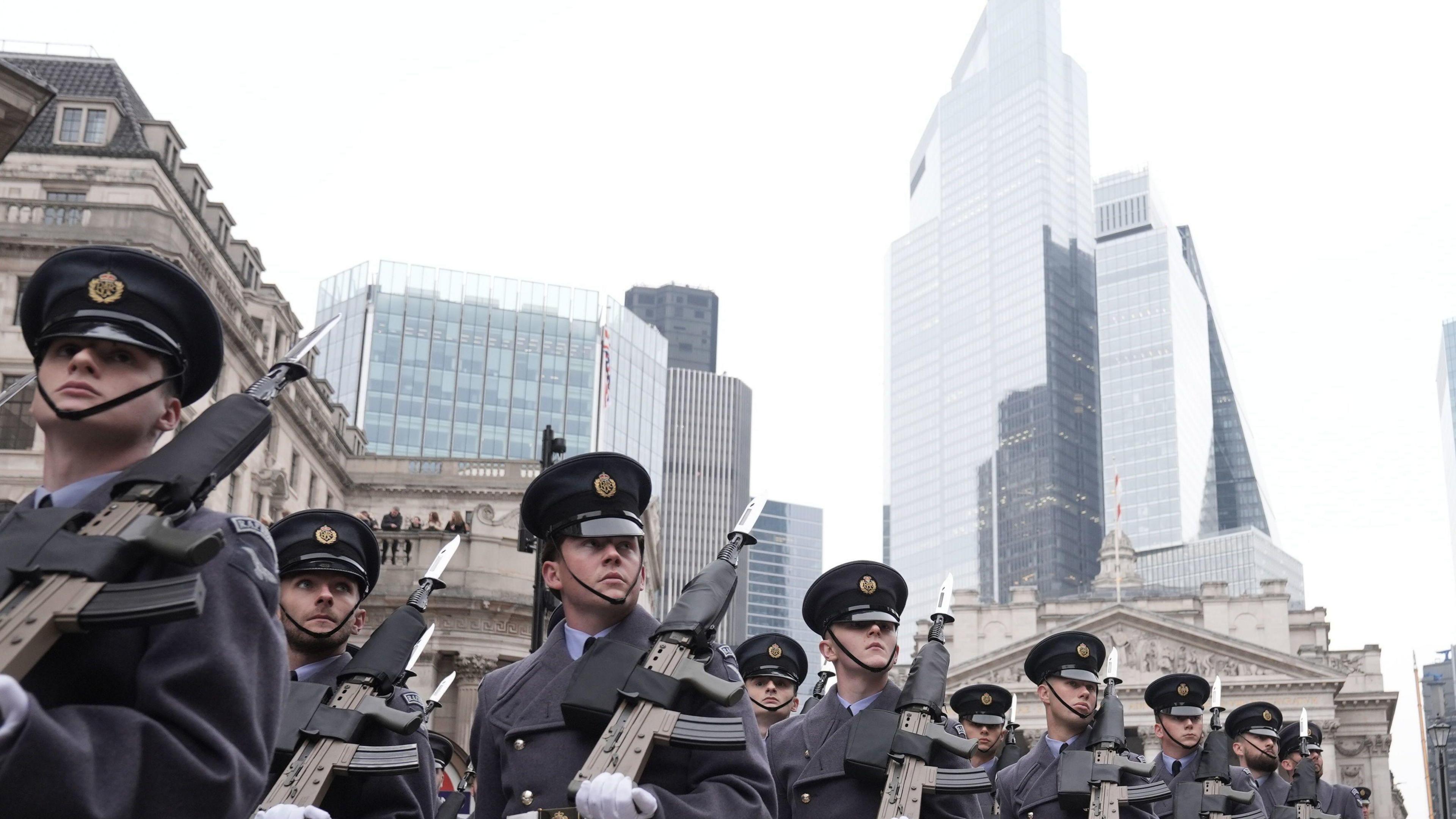 A formation of military personnel in dark uniforms with rifles march through the City of London. The soldiers are set against a background of modern skyscrapers and historic architecture.