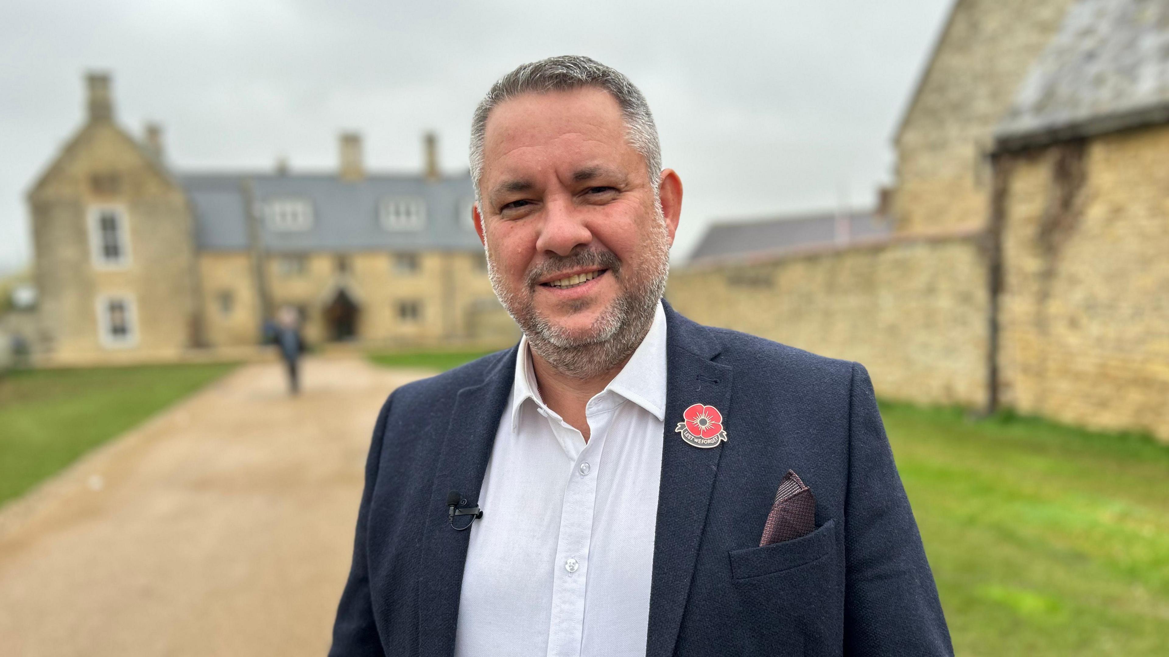 A man in a white shirt and navy blazer with a poppy badge in the lapel stands by an old building. 