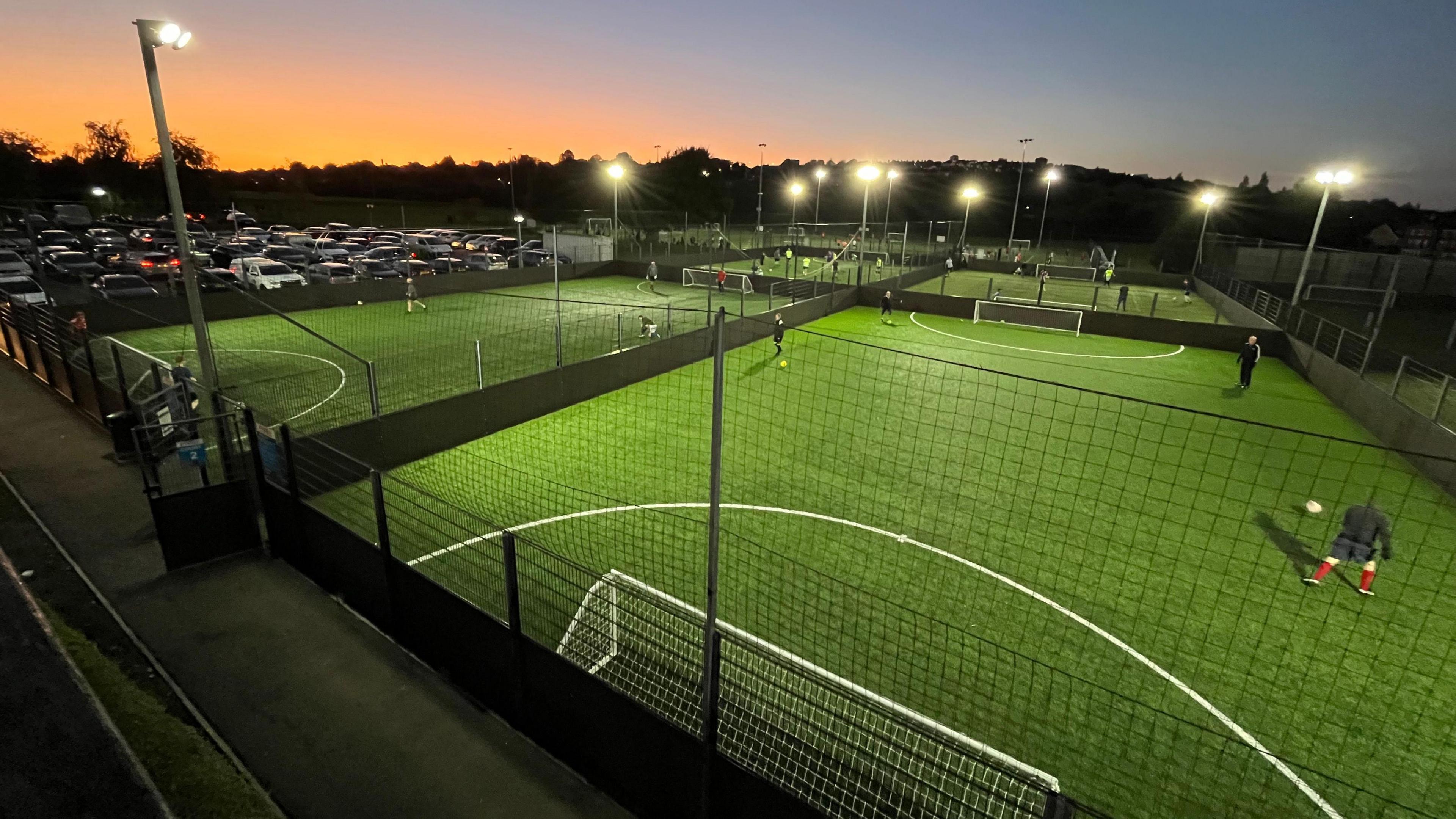 A series of five-a-side football pitches at Imperial Sports Centre in Bristol are seen from a high vantage point. The floodlights on and the distant sky has a tinge of orange as the sun goes down. There are a handful of players kicking footballs around on some of the pitches, which are covered in green astroturf.