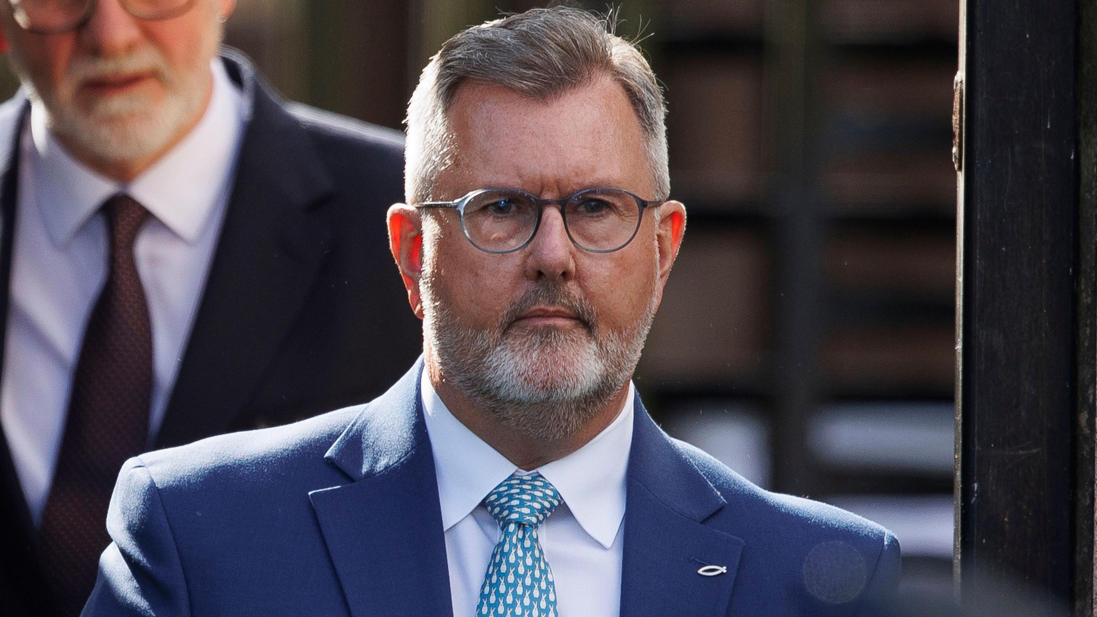 Sir Jeffrey Donaldson - a man with grey hair wearing small grey, circular glasses and a beard. He is wearing a navy suit, a white-collared shirt and a blue and white patterned tie. On the lapel is a small, ichthys symbol pin.