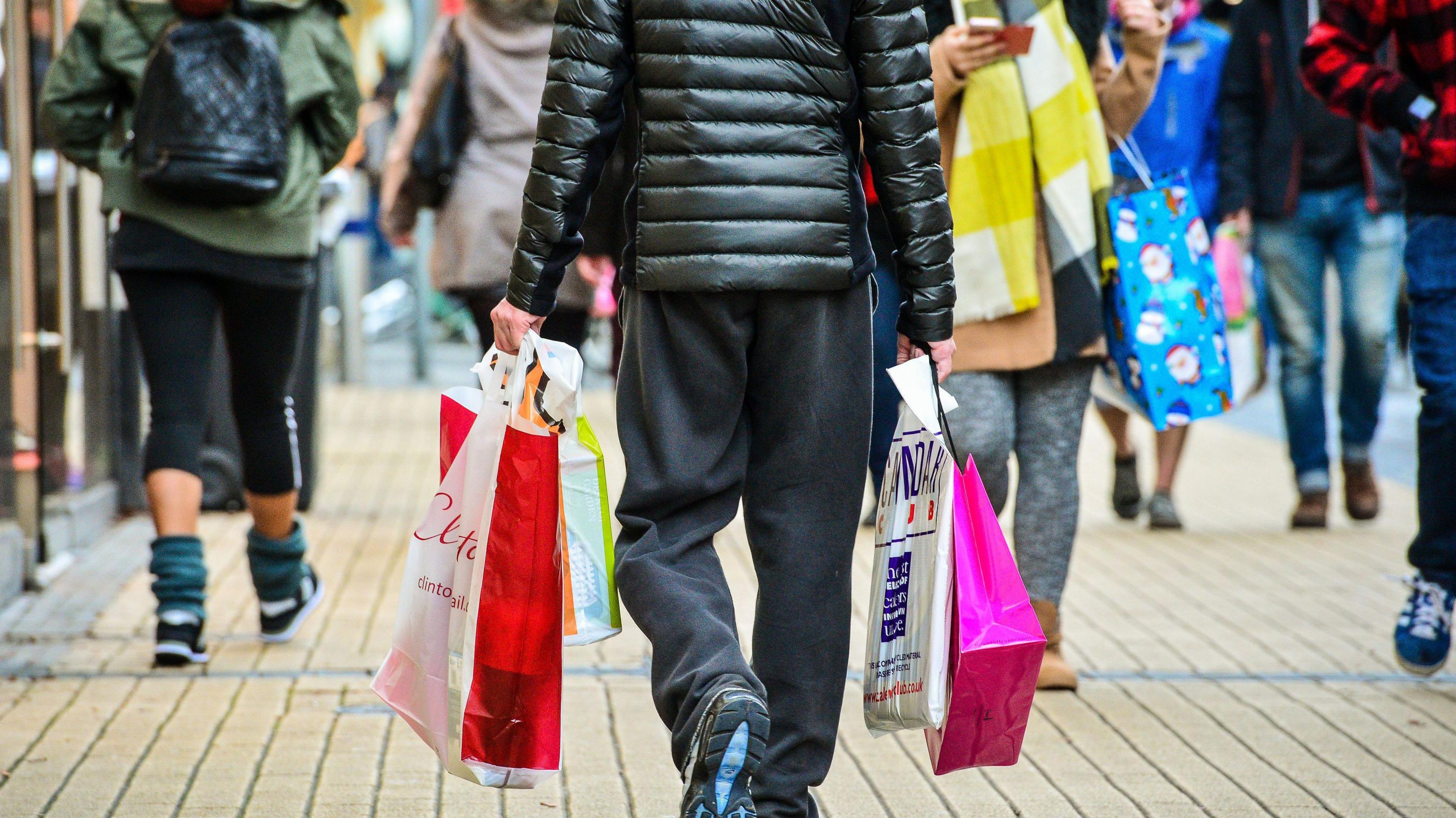 Shoppers on a street which has yellow bricks. A man carries shopping bags in both hands and wears black joggers.
