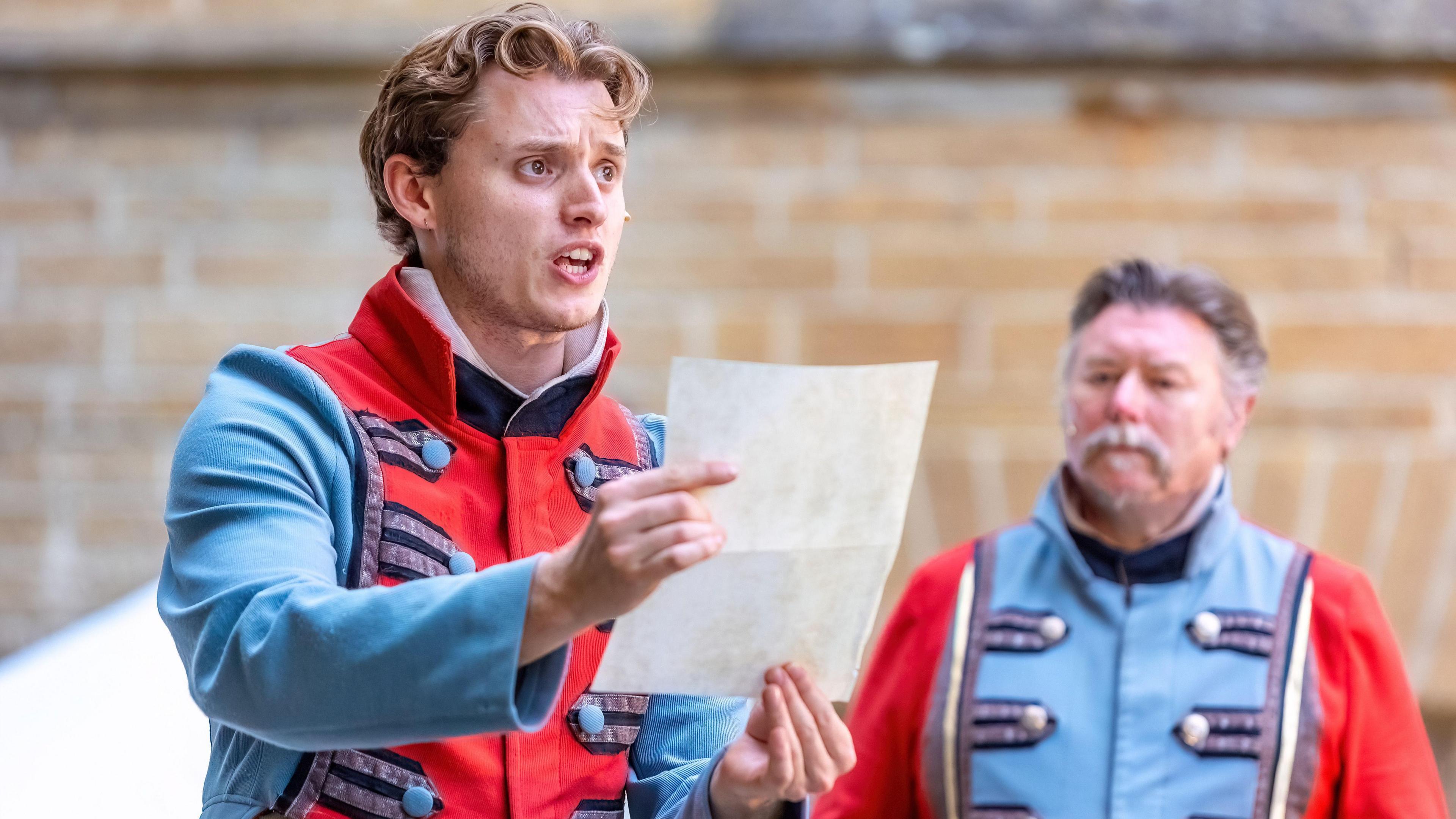 Two actors in blue and red military jackets, with the younger of the two reading from a letter he is holding up.