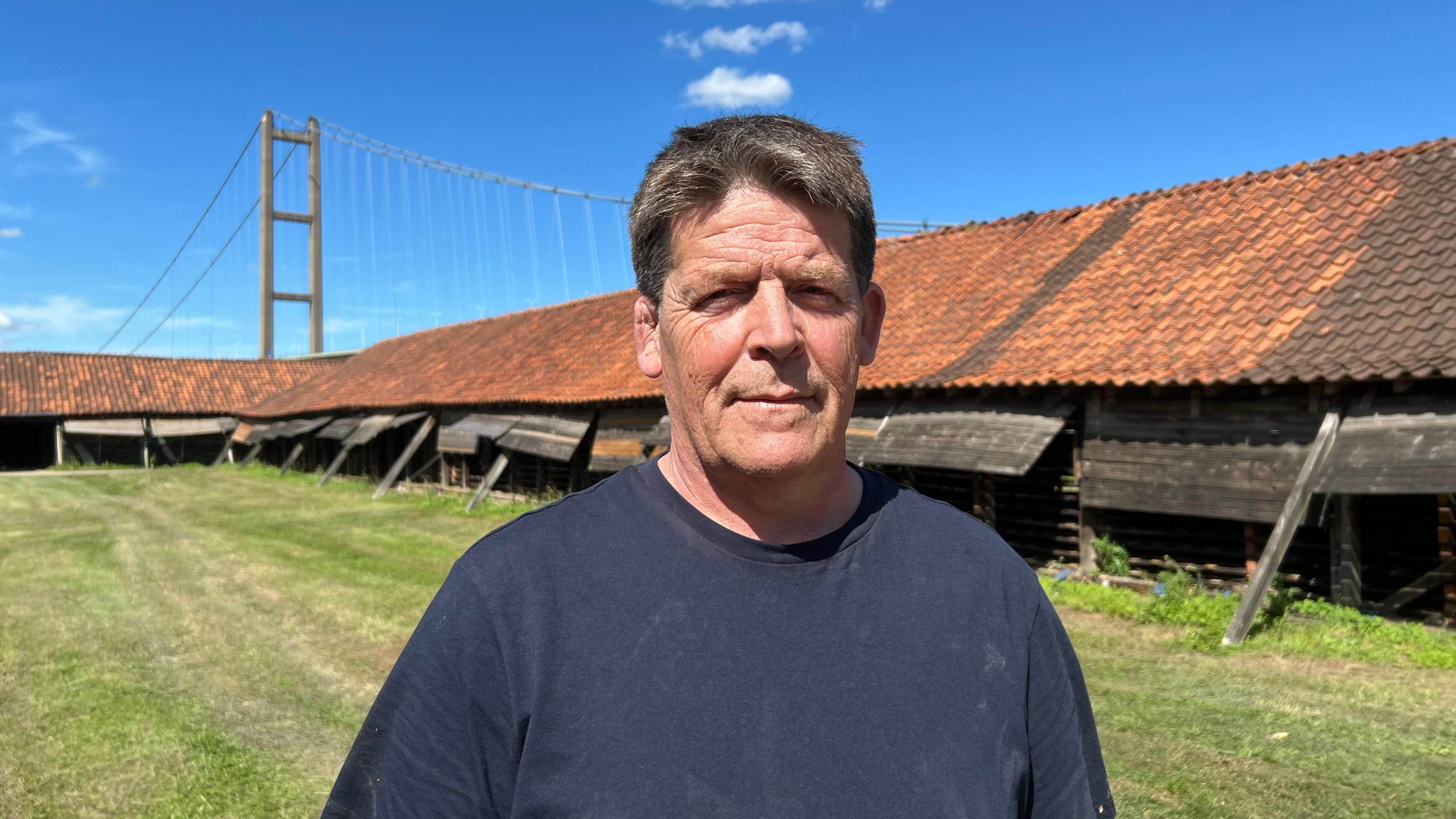 Gabriel Nichols wearing a dark navy T-shirt standing outdoors on a green lawn in front of a long rustic wooden building with a red-tiled roof. Behind the building to the left, a large suspension bridge - Humber Bridge - rises against a bright blue sky with a few white clouds. The image is brightly lit by natural sunlight.