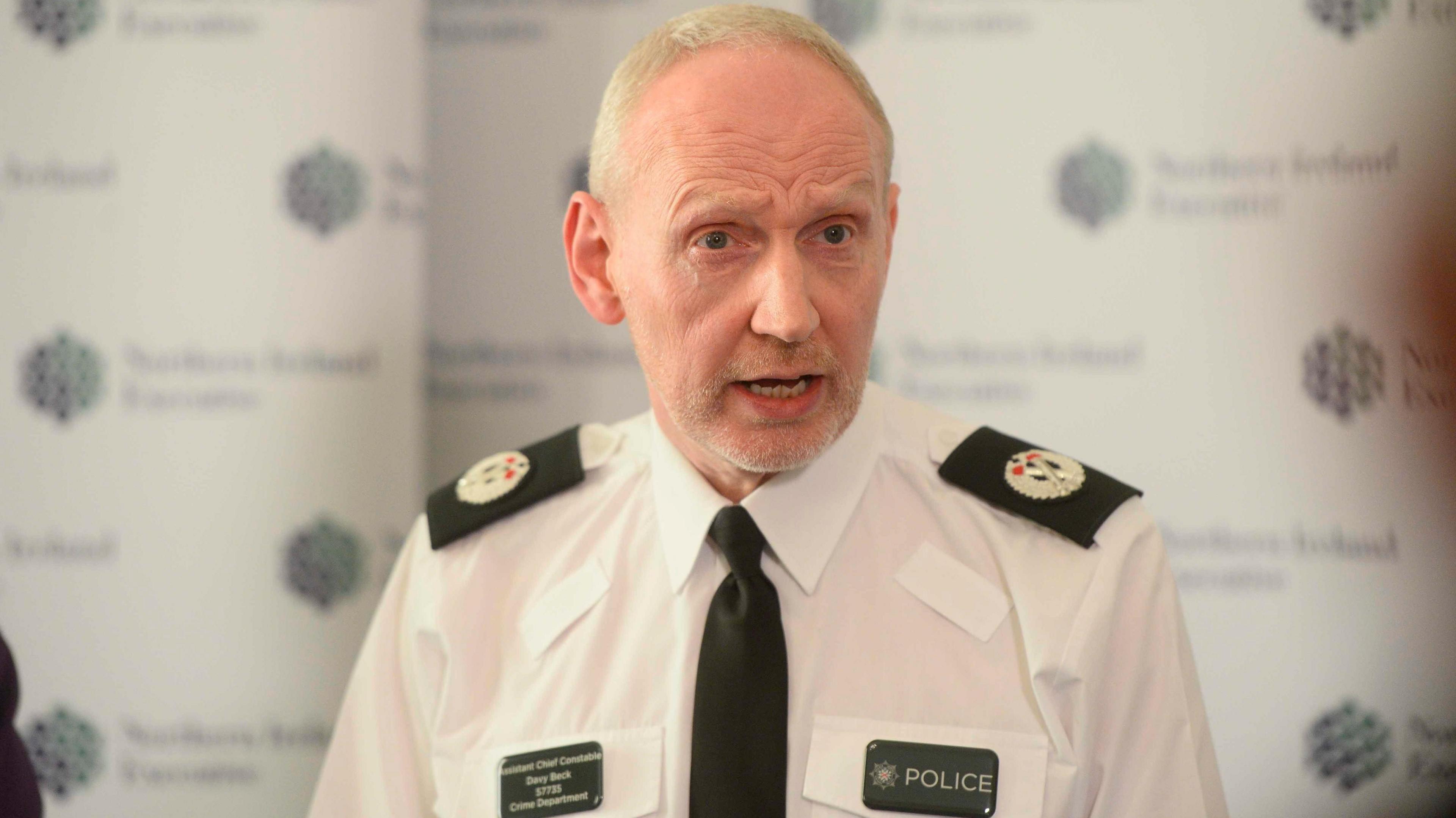 A man dressed in a police uniform consisting of a white shirt, black tie and name badge looks to the side of the camera. He has greying hair. 