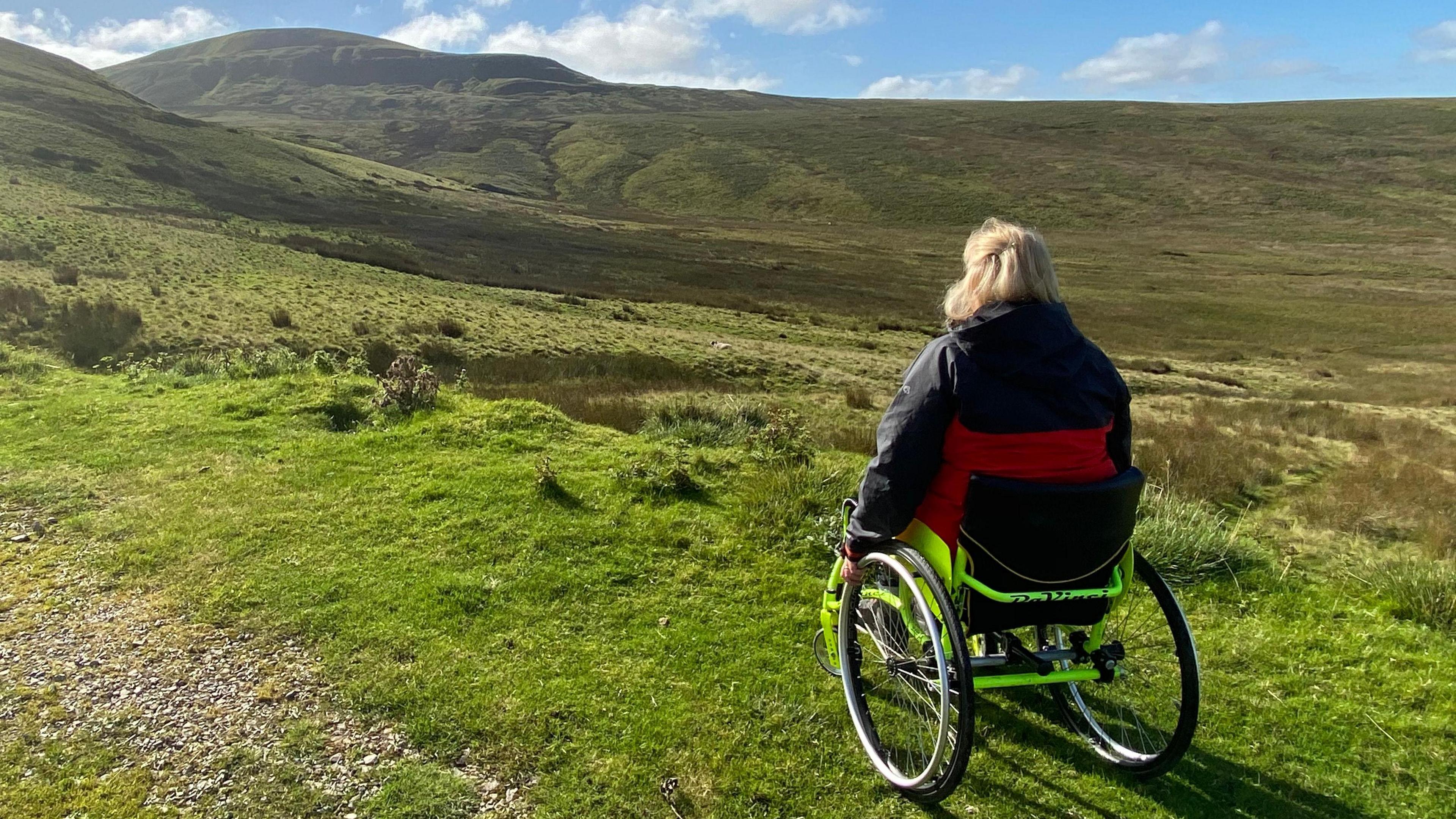 A woman sitting in a wheelchair wearing a black coat is looking out across lush green moorland.