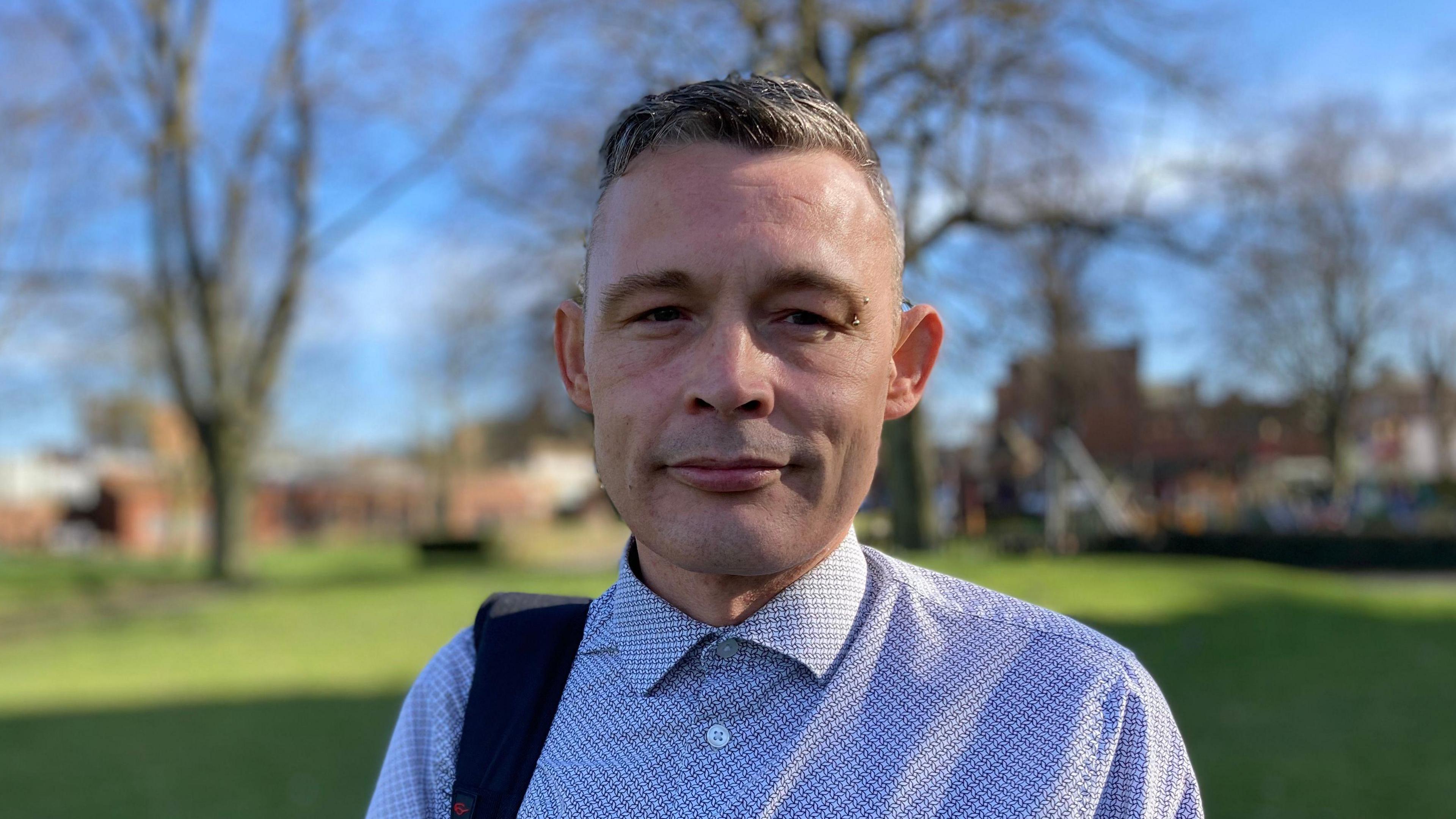 A man with short grey hair and wearing a blue patterned shirt with a piercing above his left eye. He is standing in a field and looking into the camera.