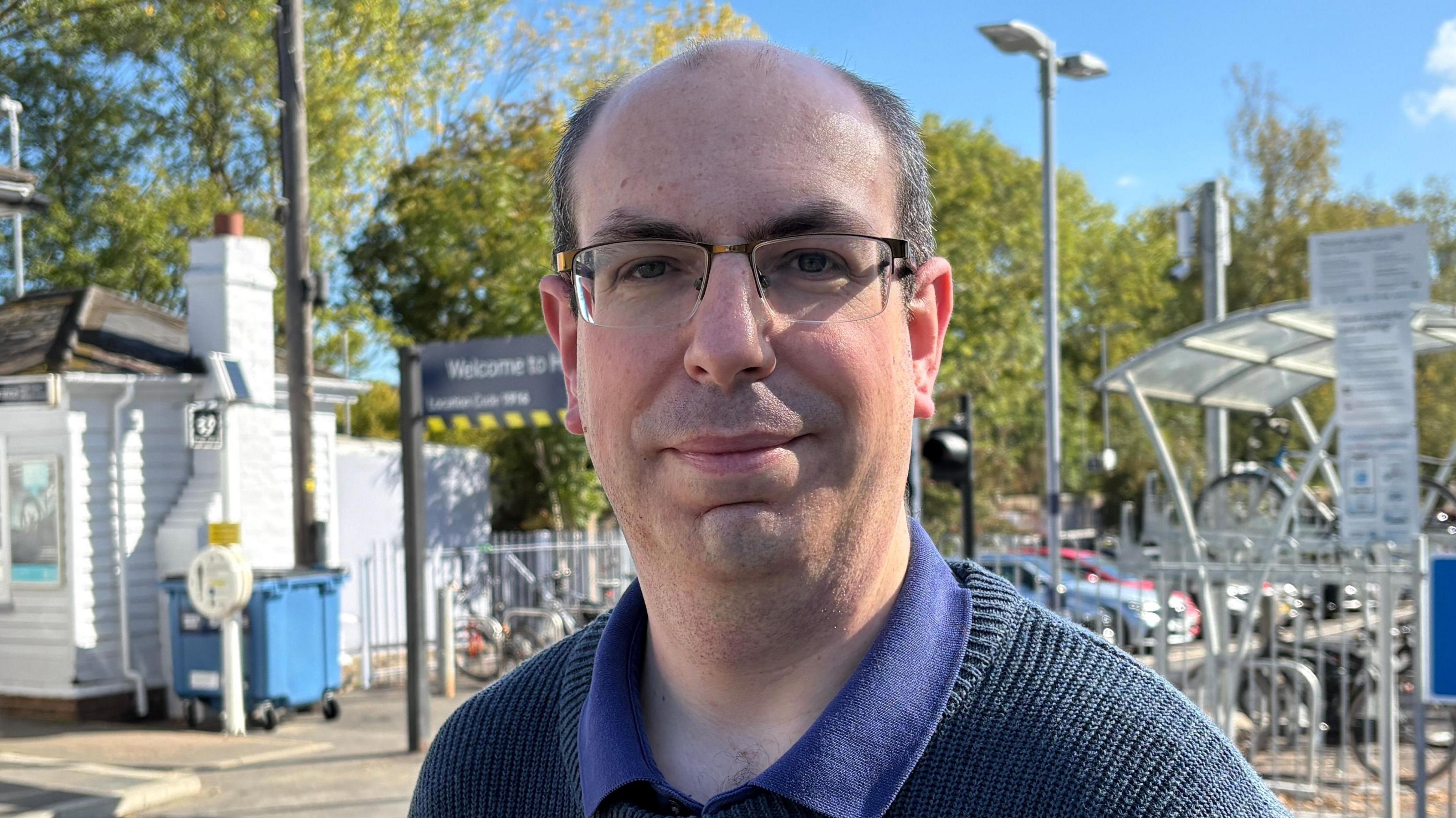 Rob Mansfield looking directly down the camera. He is wearing a dark coloured knitted jumper over a blue, collared t-shirt. He has rectangular framed glasses on and is smiling a little, though not showing his teeth. He is bald on the top of his head and has dark hair on the sides. He is standing outside and in front of a car park. A low-rise, white building is on the left of the picture and there is a bike rack and some bikes on the right of frame. Further back are some mature trees. It is a sunny day and there is just one small cloud in an otherwise blue sky.