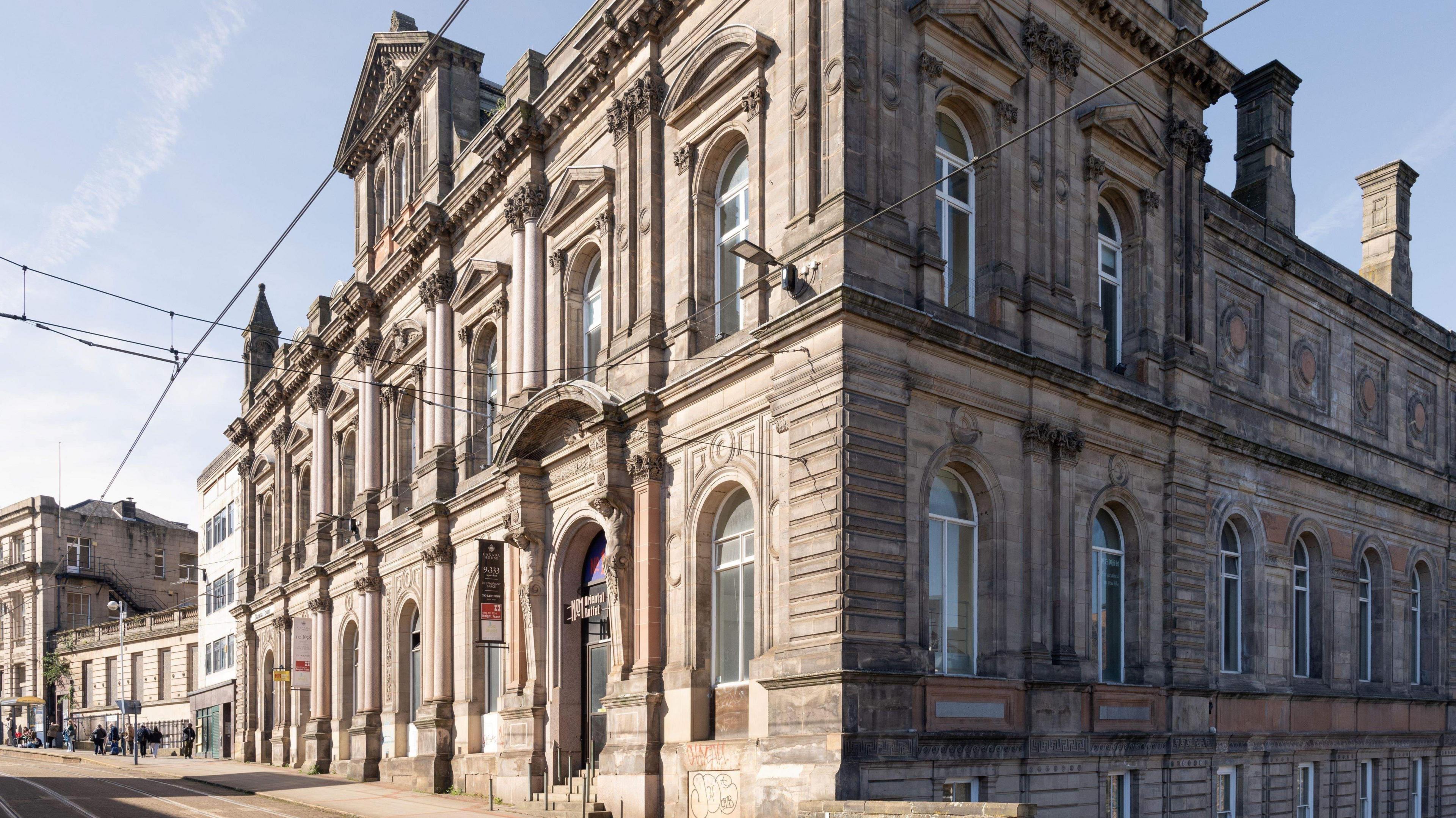 A large stone built building with tall arched windows, columns and decorative stonework stands on the corner of a street