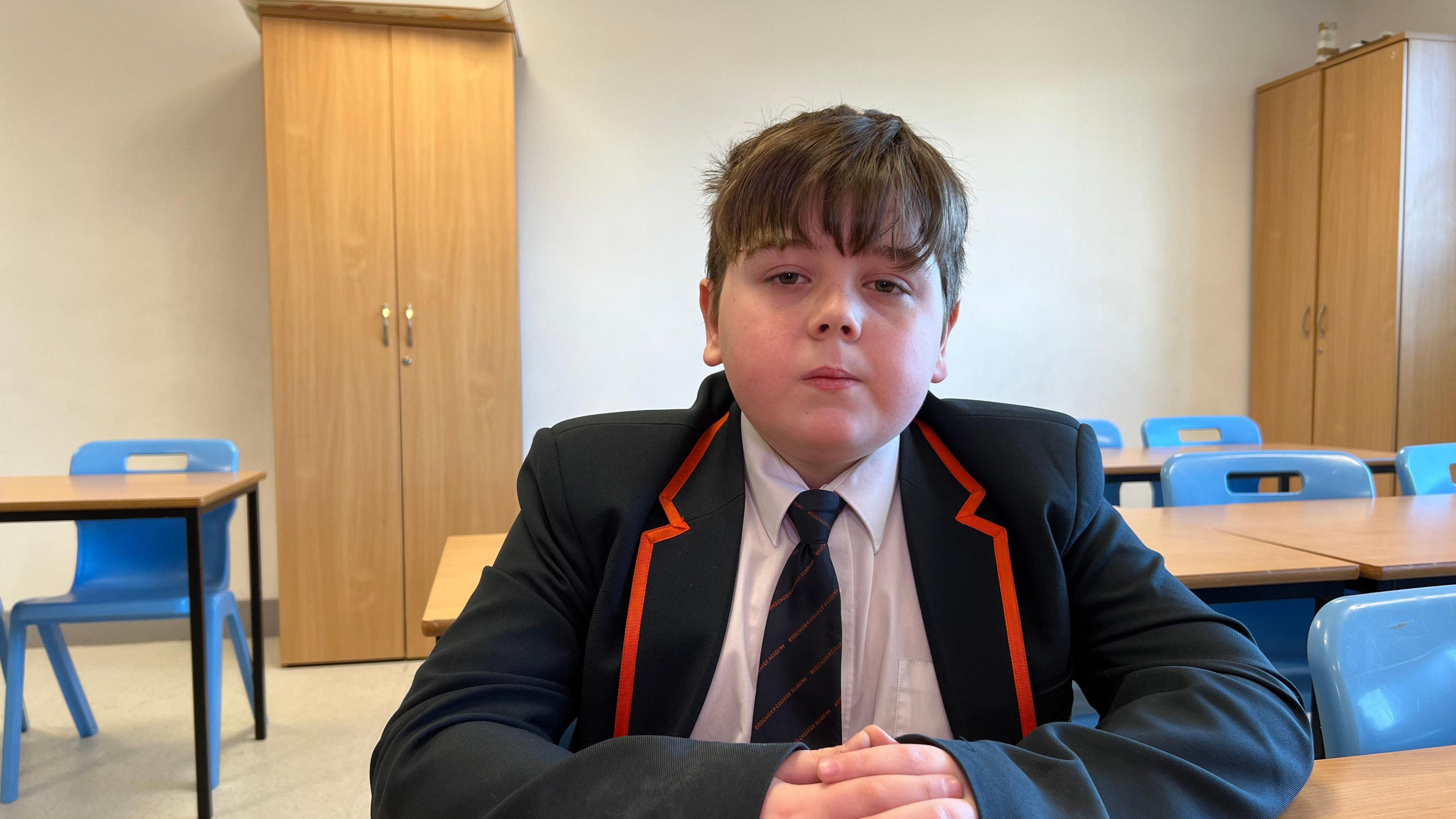 A male pupil in a dark blazer, white shirt and dark tie looks at the camera. He has short hair and is sitting in an empty classroom